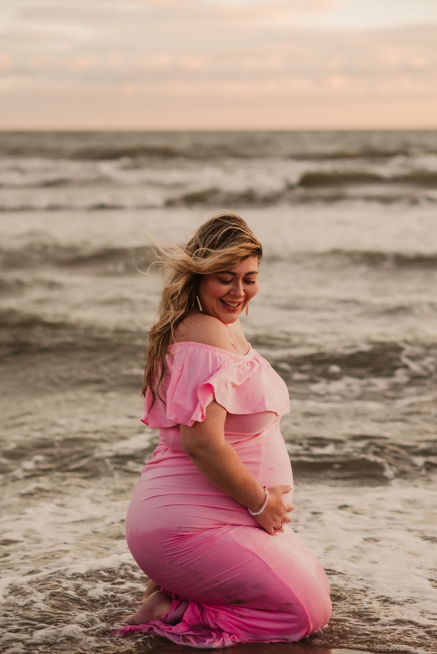 A person in a pink dress sits at the beach during sunset while waves crash behind them.