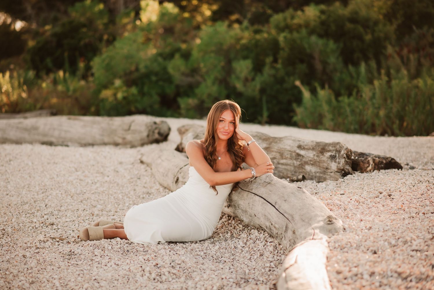 A person in a white dress sits gracefully on a rocky beach during golden hour sunset.