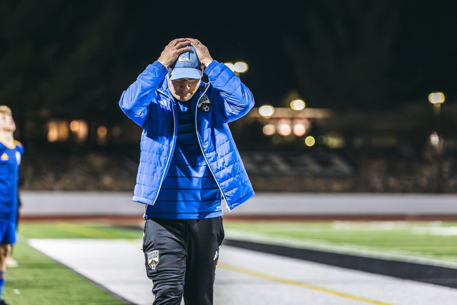 A person in a bright blue winter coat stands on a sports field at night with stadium lights glowing in the background.