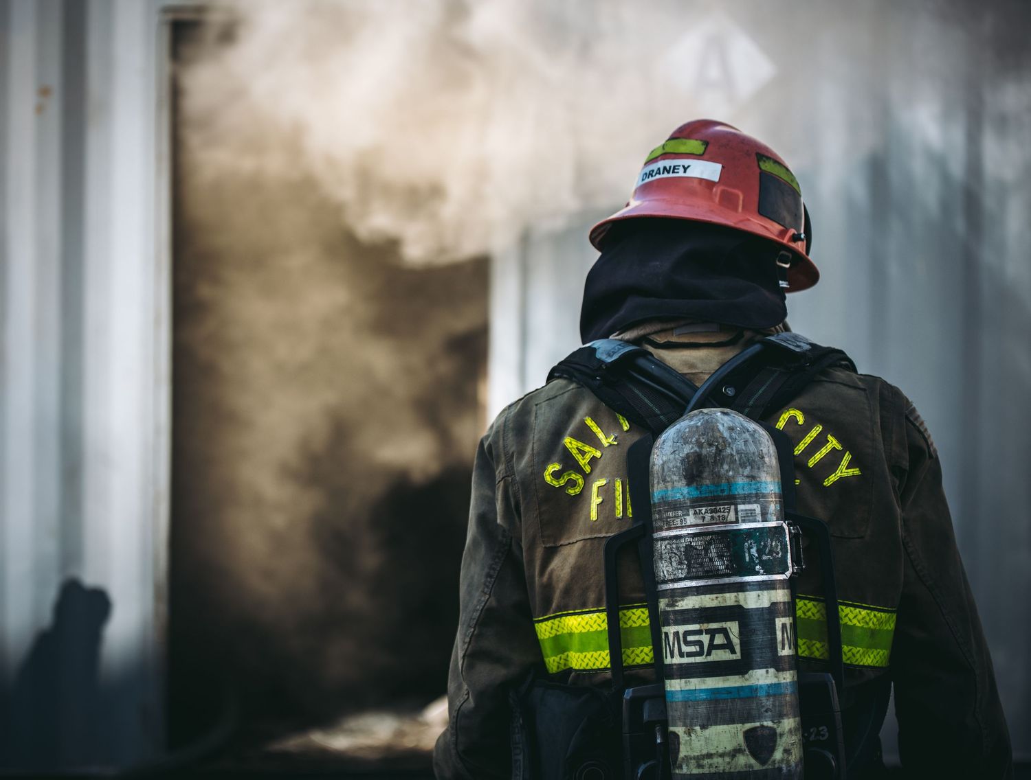 Firefighter wearing protective equipment and oxygen tank stands amid smoke during emergency response.