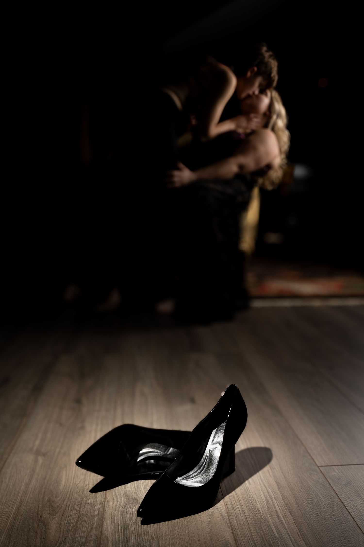 Black high heels cast dramatic shadows on wooden floor in low-key lighting.
