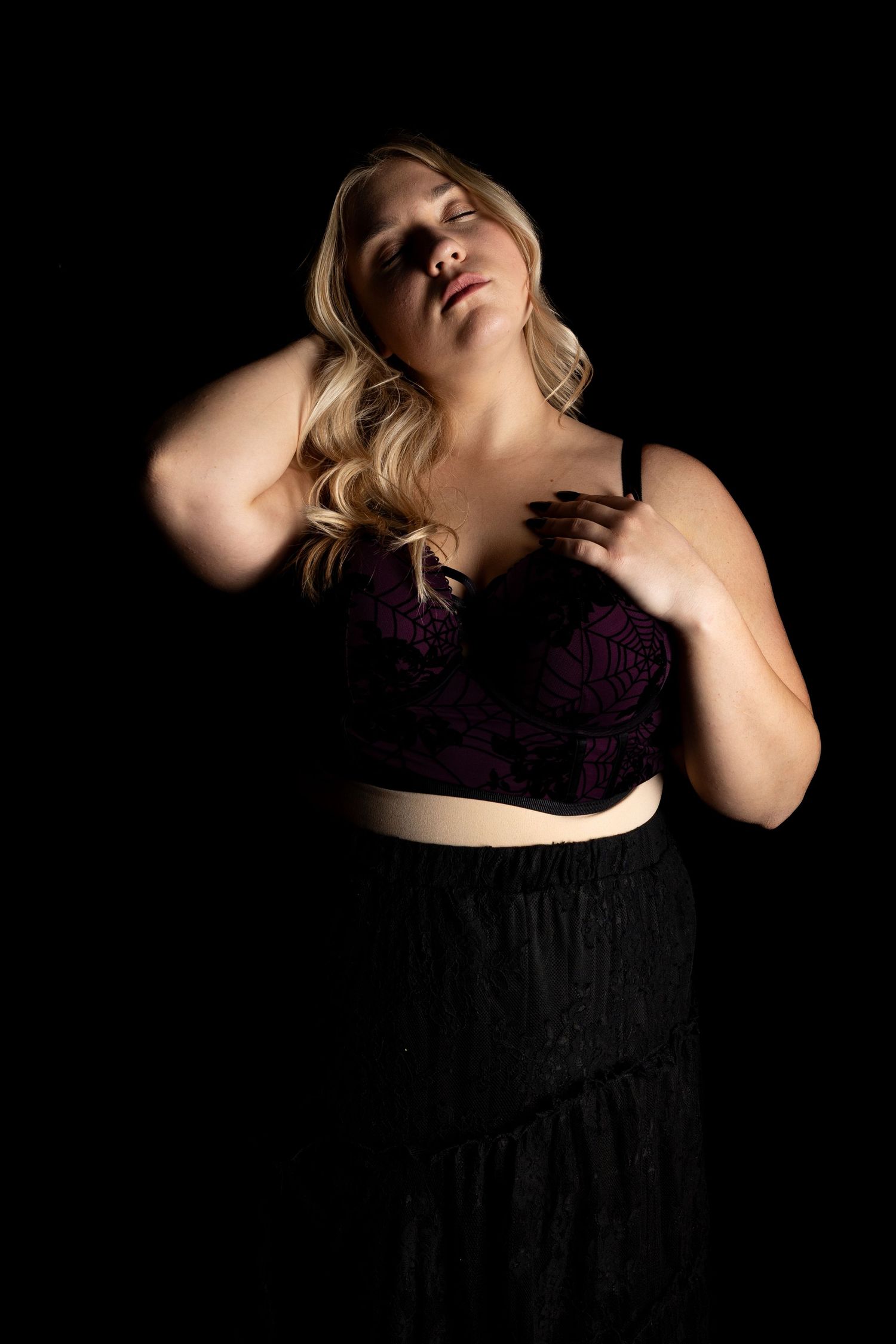 Artistic boudoir portrait in dark lighting with purple lingerie against black background.