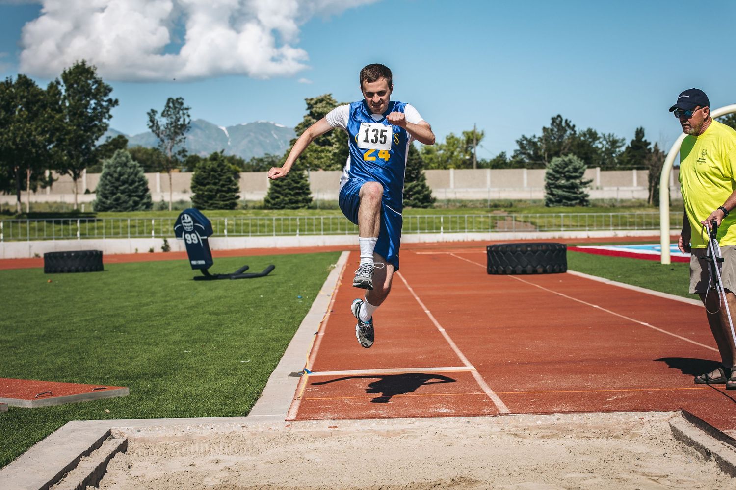 Athletes compete in long jump events at an outdoor track and field competition on a sunny day.