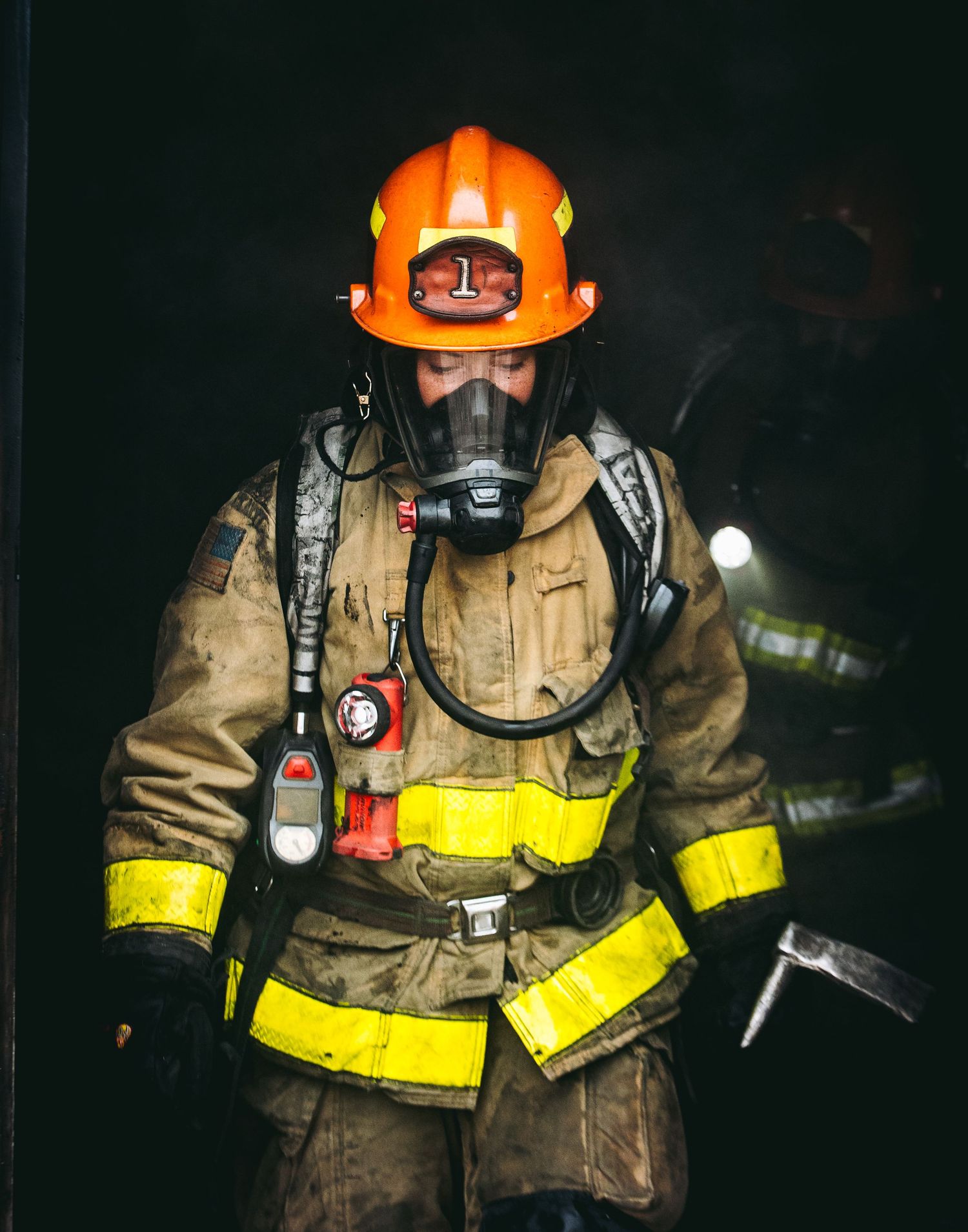 A firefighter in full protective gear with orange helmet and breathing apparatus stands ready in dark surroundings.