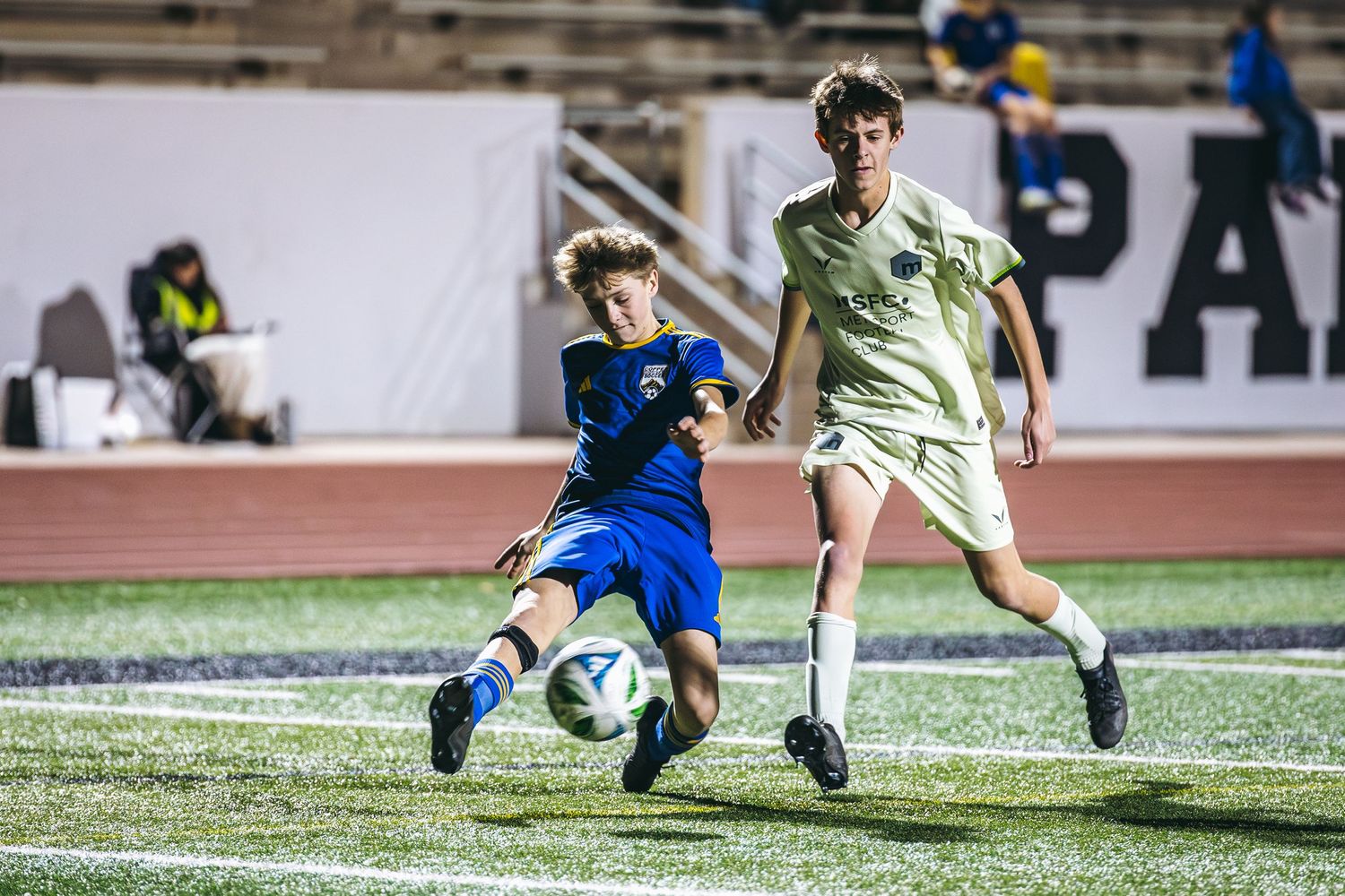 Two soccer players compete for the ball during a match on an artificial turf field under stadium lights.