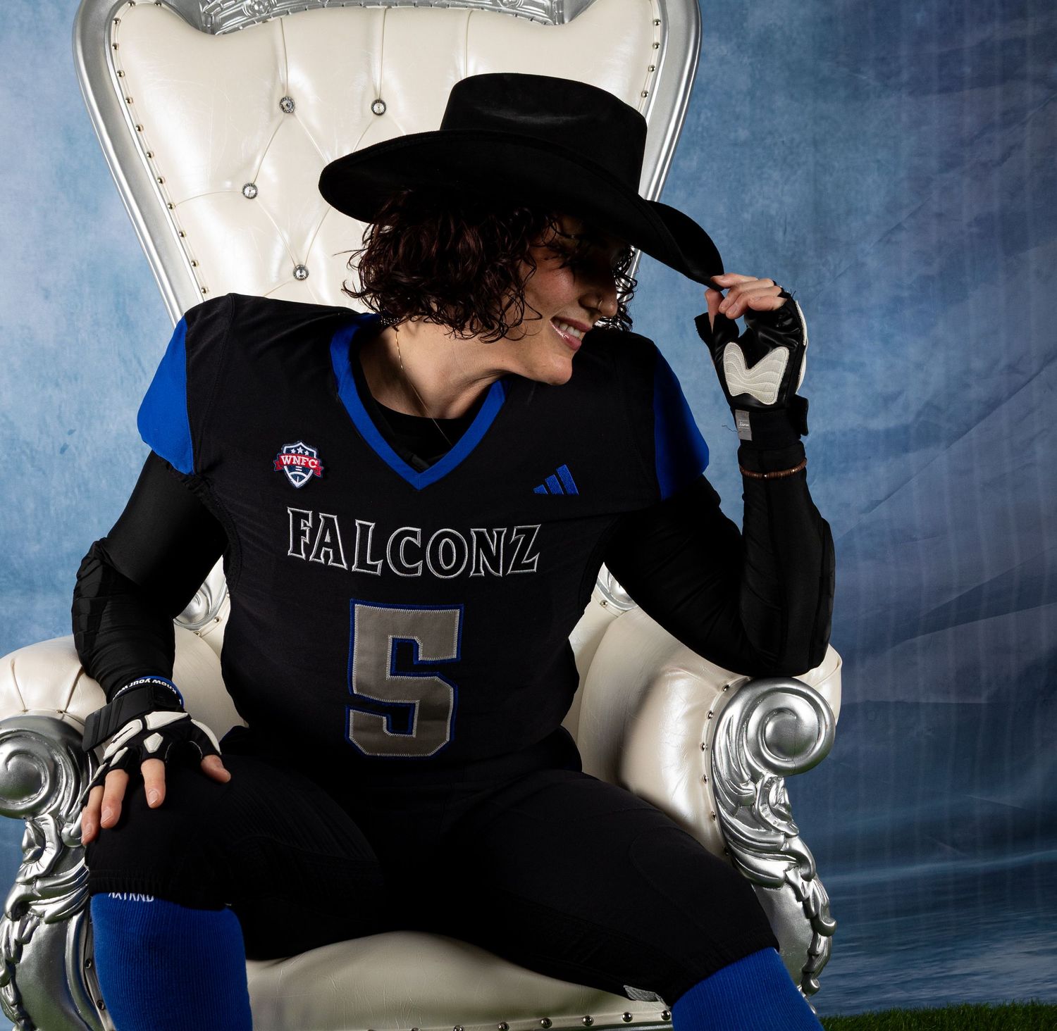Football player in black and blue uniform sits in contemplative pose against dramatic blue backdrop.