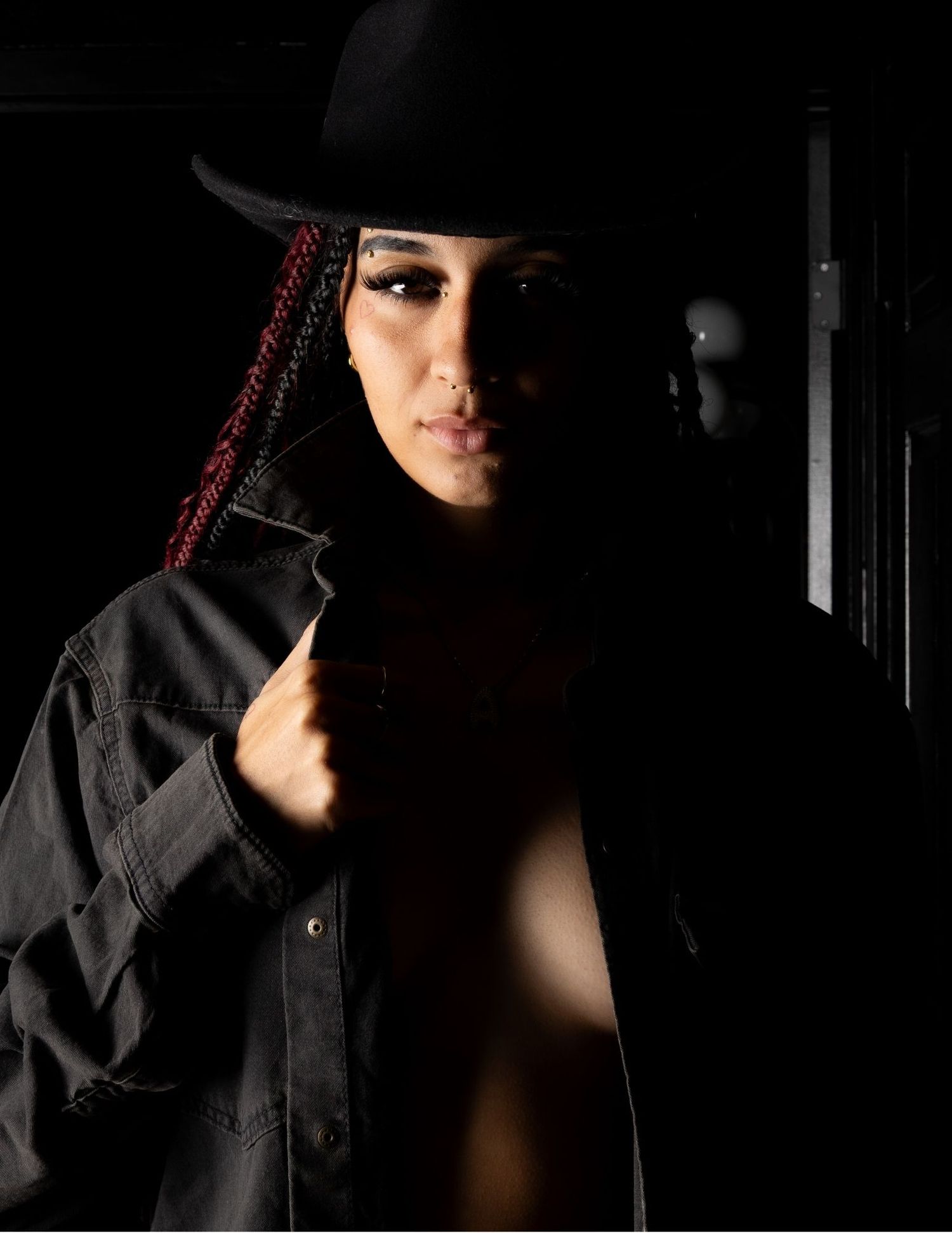 A dramatic low-key portrait in black and white showing a figure wearing a dark hooded jacket against a shadowy background.