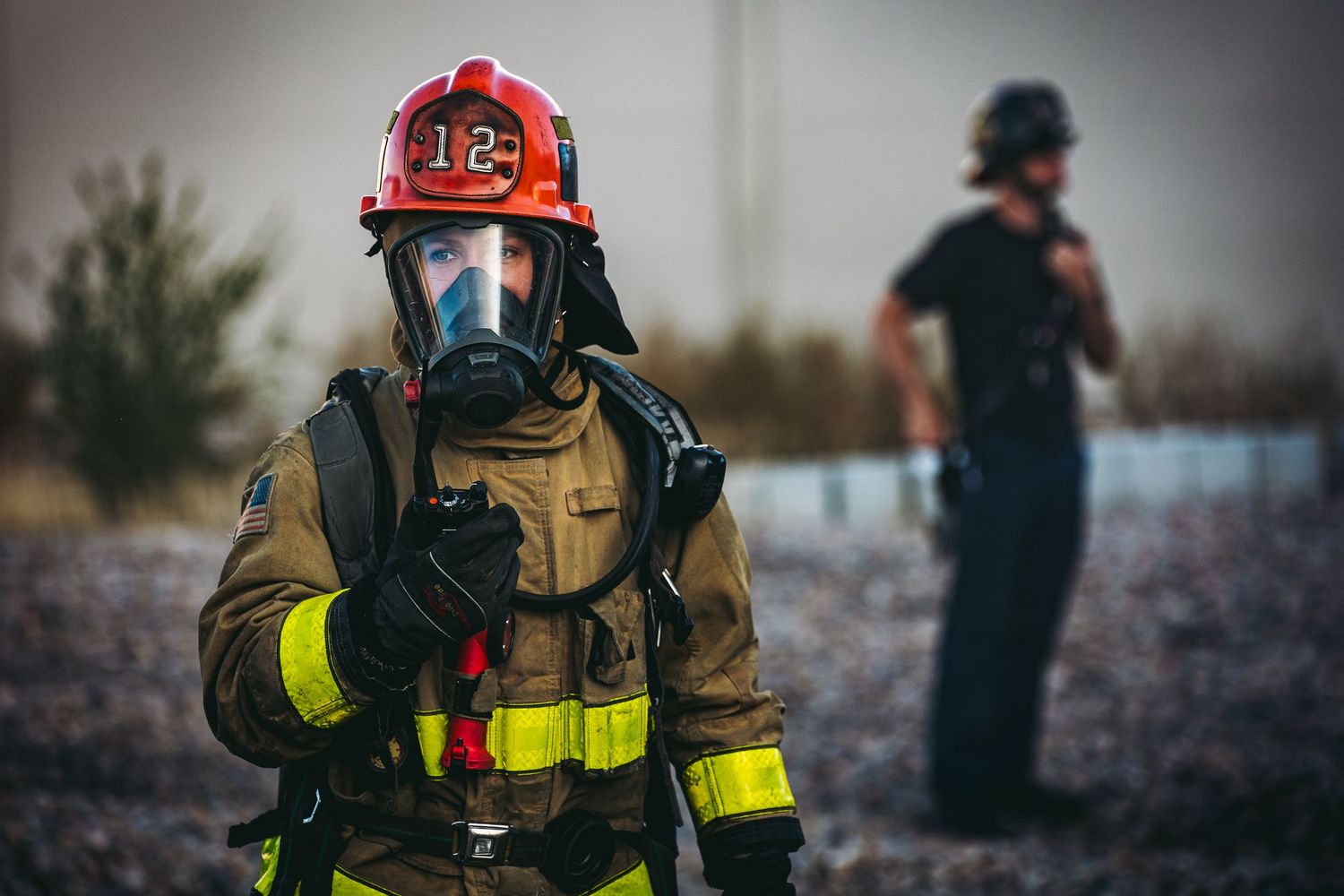 Firefighter in full protective gear with red helmet and breathing apparatus stands while another figure is visible in background.