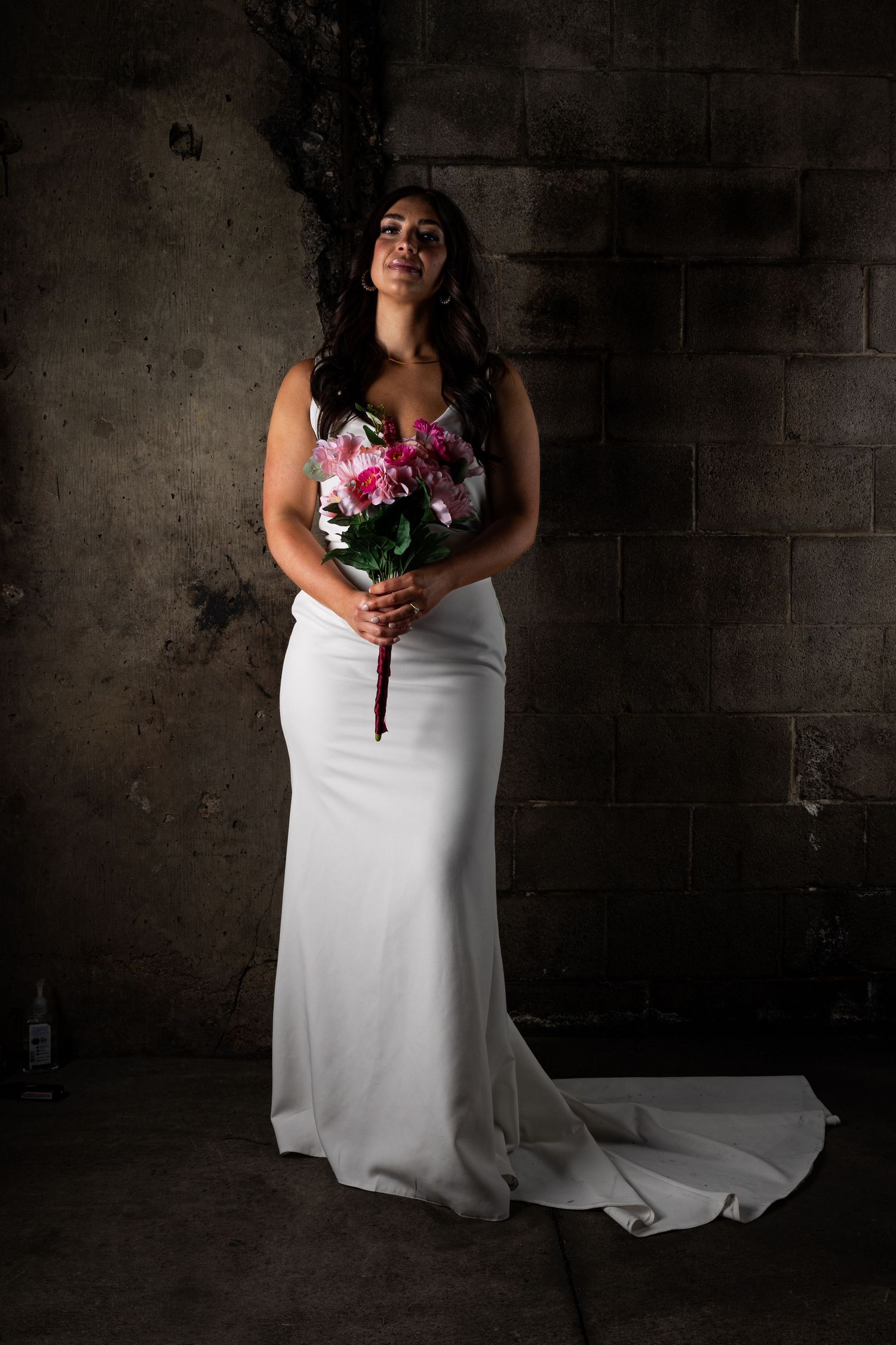 Person in white wedding dress holding colorful bouquet against dark wall.