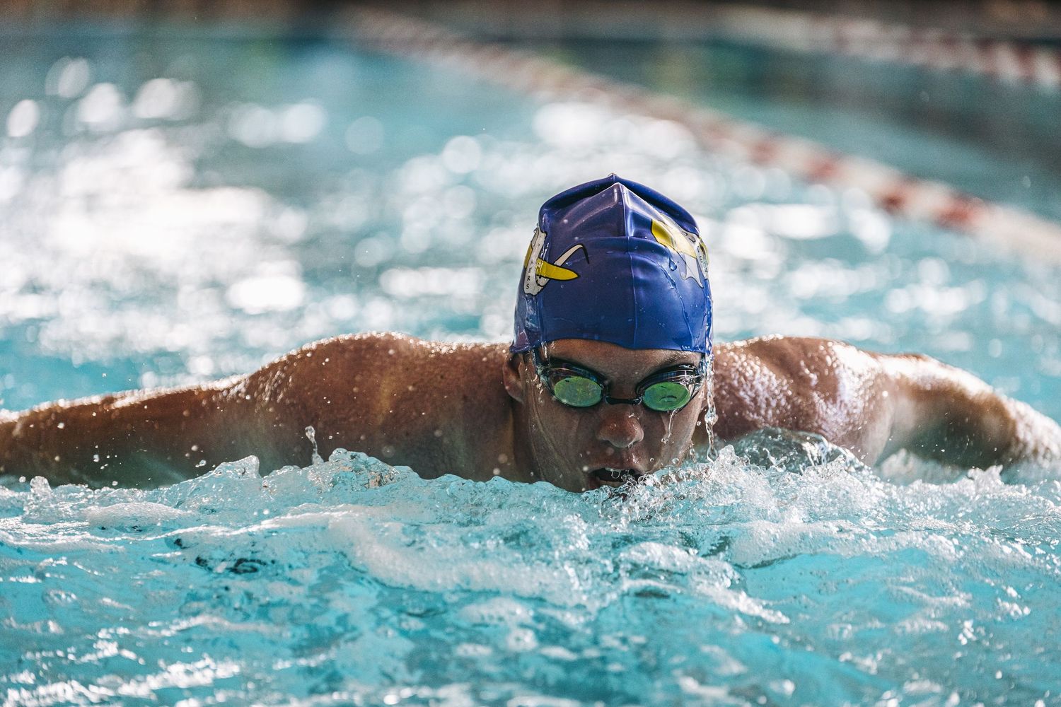 Competitive swimmer performs butterfly stroke in indoor pool wearing blue swim cap and goggles during training.