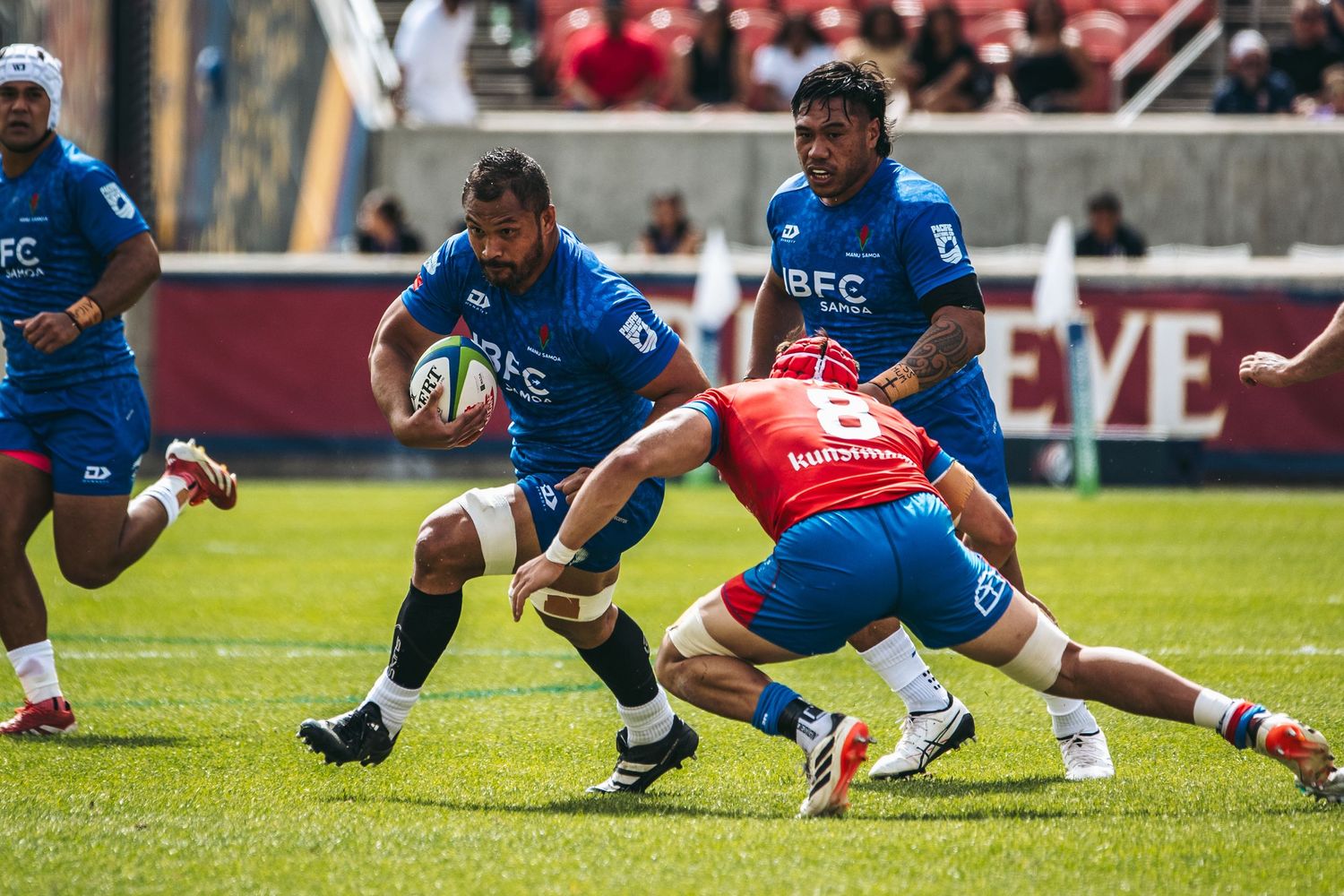 Rugby players dive and stretch during an intense match between teams in red and blue uniforms on a grass field.