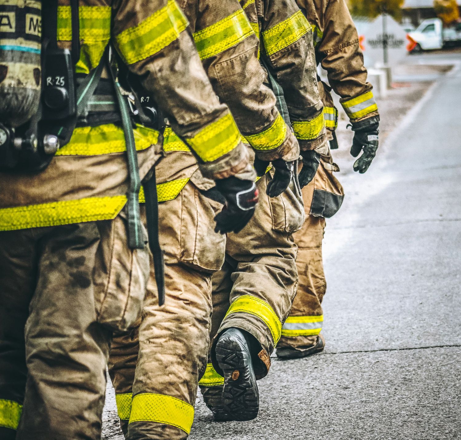 Firefighters in tan turnout gear and yellow reflective stripes stand in formation on a paved surface.