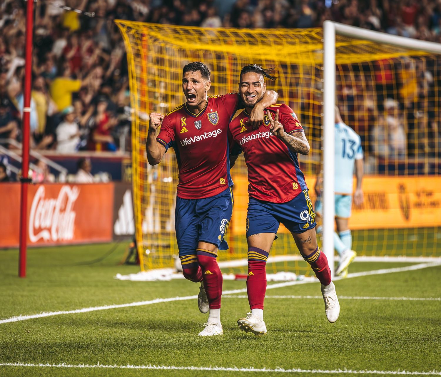 Soccer players celebrate a goal during a match with yellow goal posts and netting visible in the background.