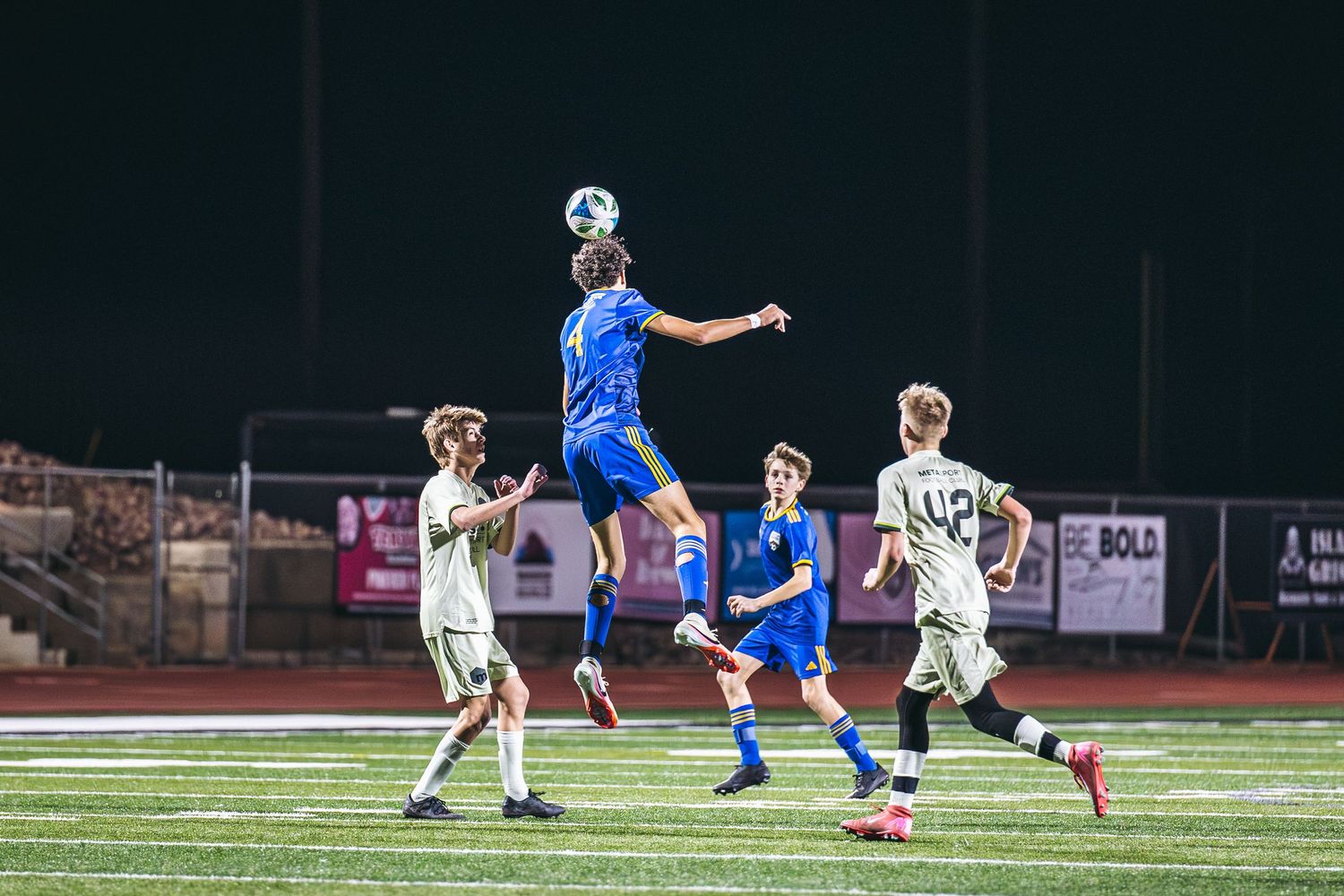 Soccer player in blue jersey jumps high to head the ball during competitive match against players in white uniforms.