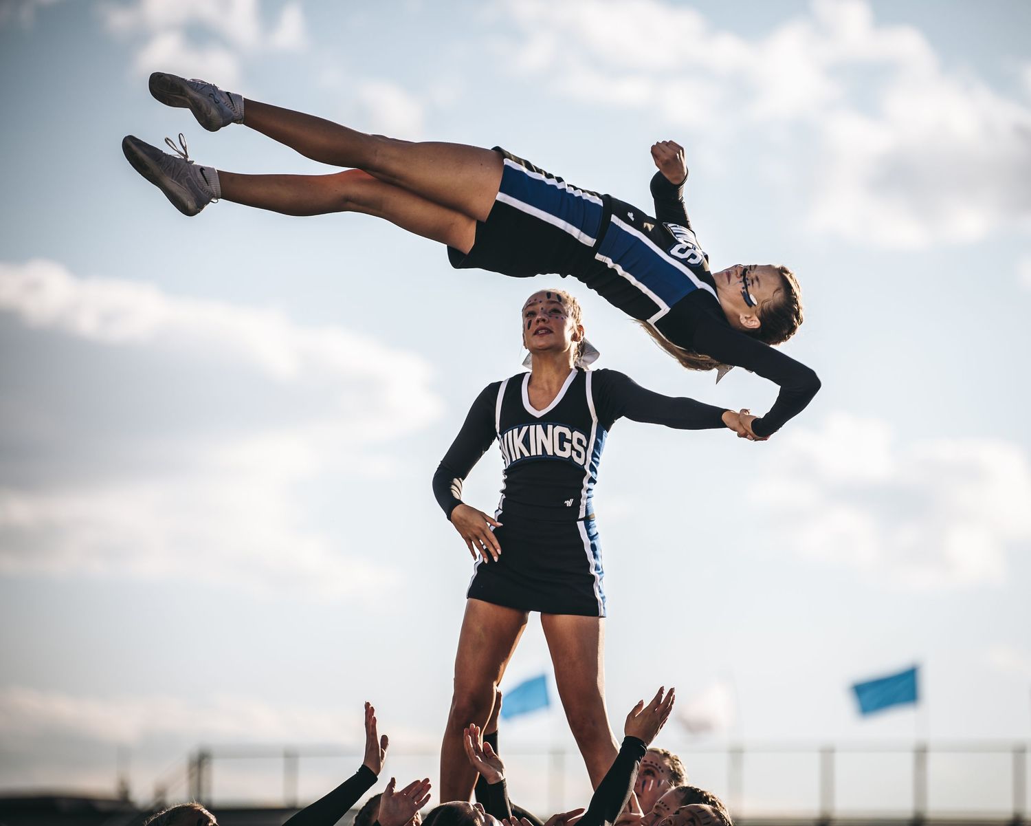 Cheerleaders performing a stunt sequence with lifts and tosses against a cloudy sky backdrop.