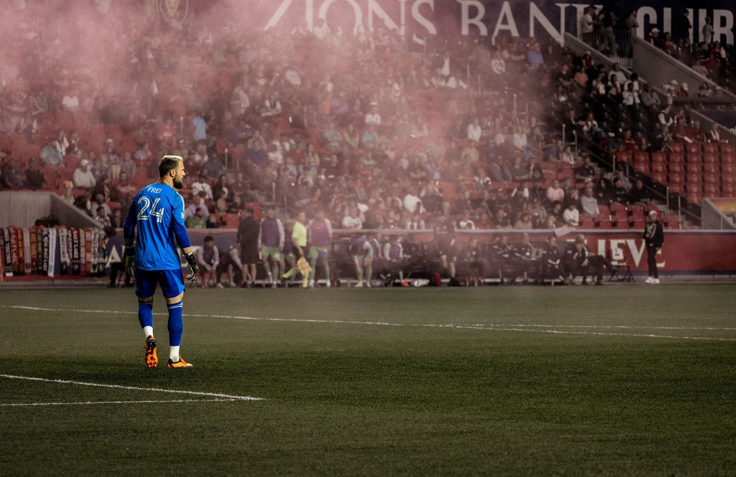 Soccer player in blue uniform stands on field while red smoke fills the stadium atmosphere during match.