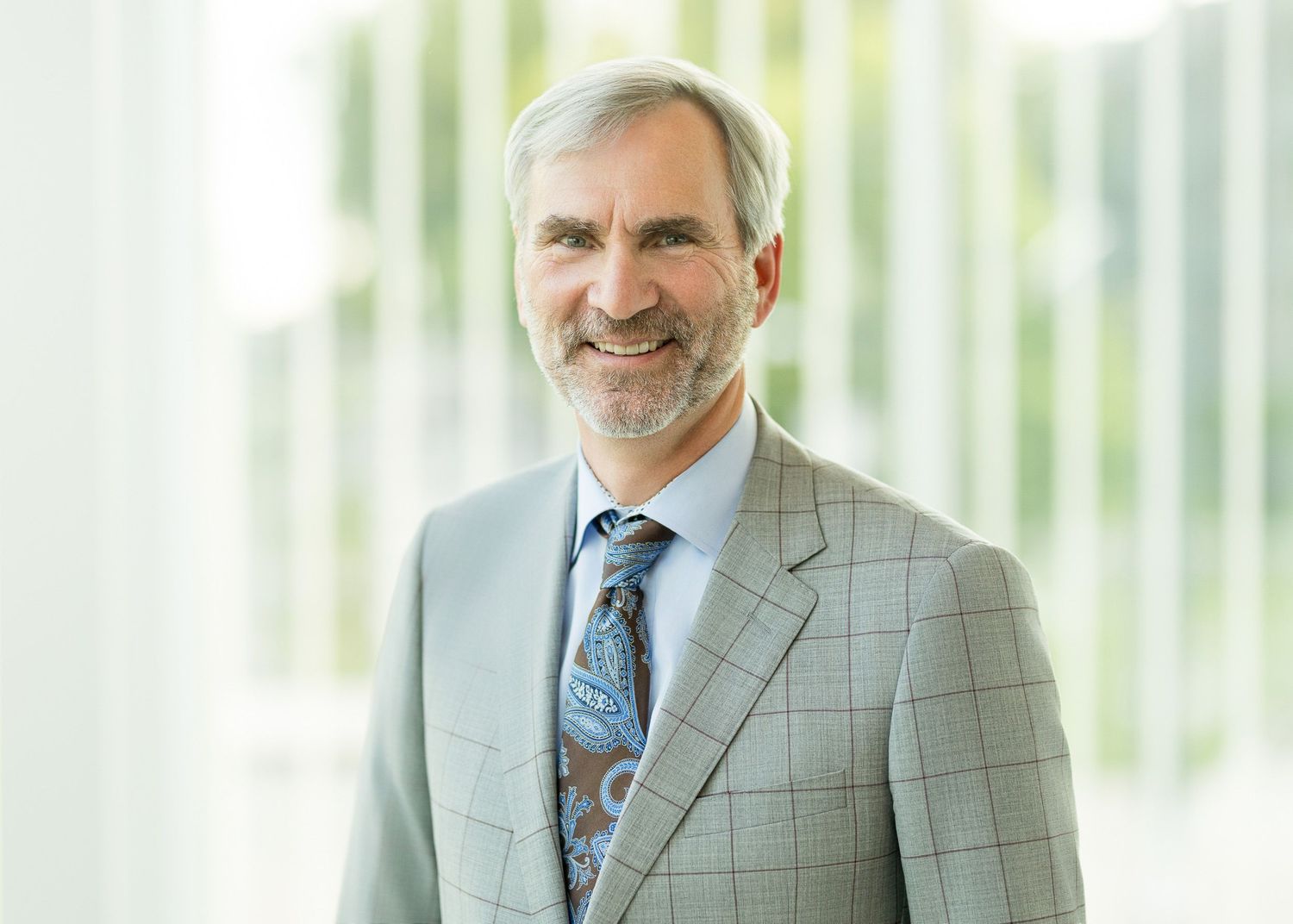 Professional headshot of a smiling middle-aged man in a gray suit and patterned tie against a bright modern background.