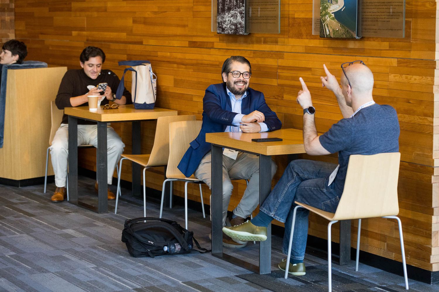 Three men sit at tables inside the Vancouver Convention Centre, two conversing while one uses a smartphone.