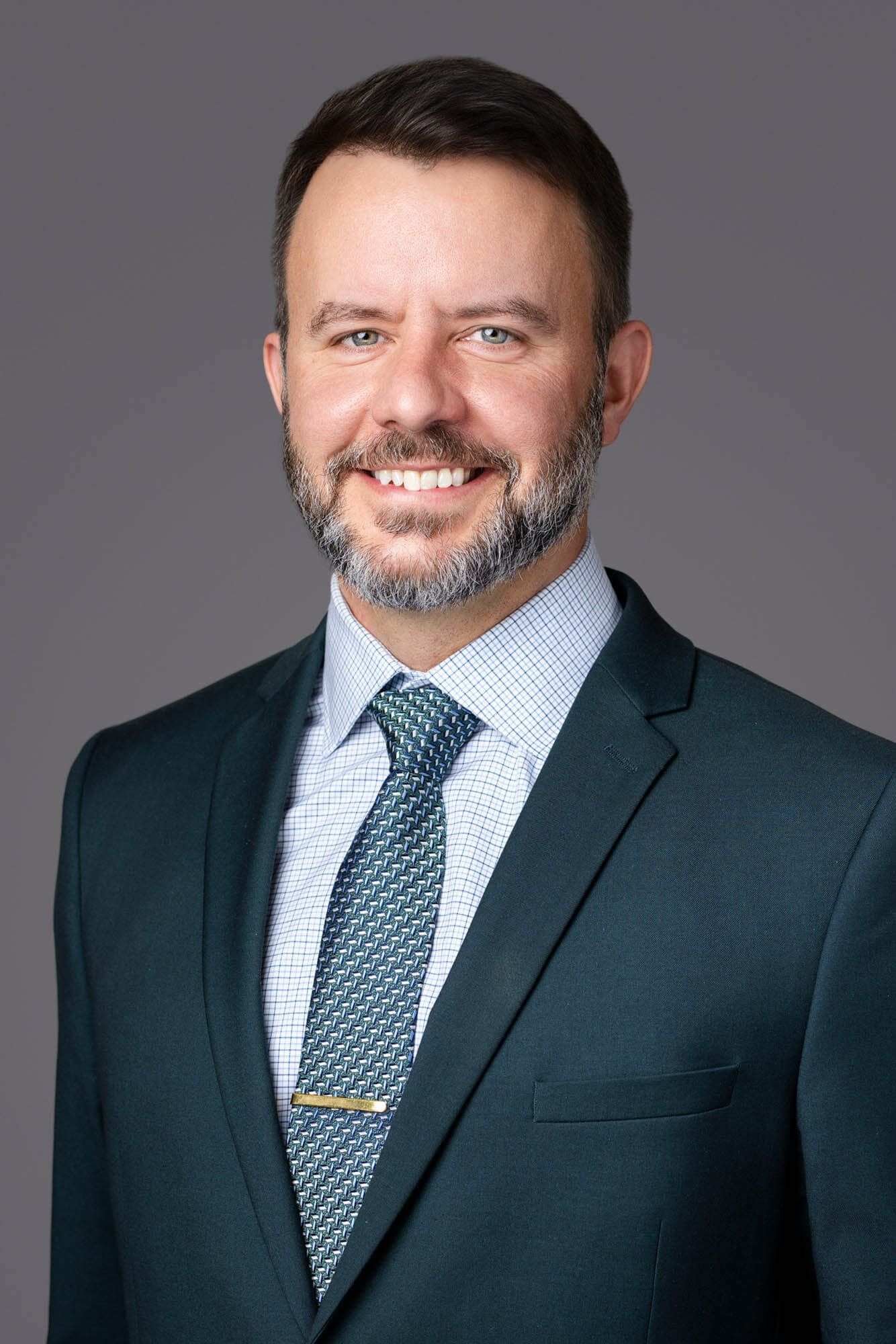 Professional headshot of a smiling man with beard wearing dark green suit and patterned tie against gray background.