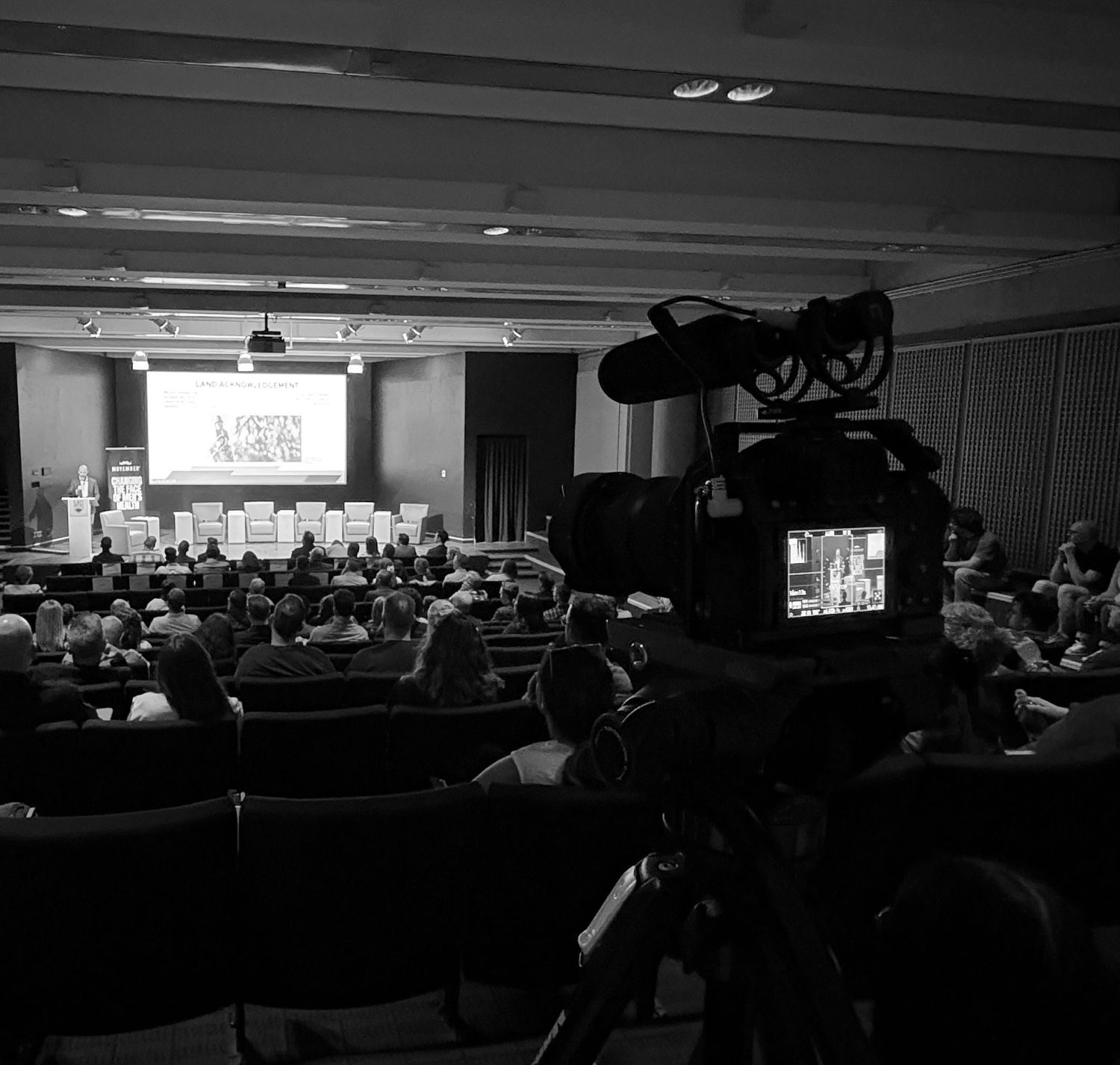 Black and white photo of a video camera filming a conference presentation in a packed auditorium with a lit screen on stage.