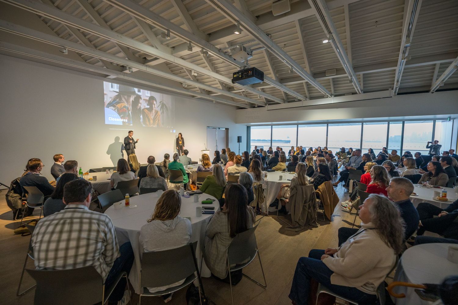Large conference room filled with attendees seated at round tables listening to speakers presenting on stage.