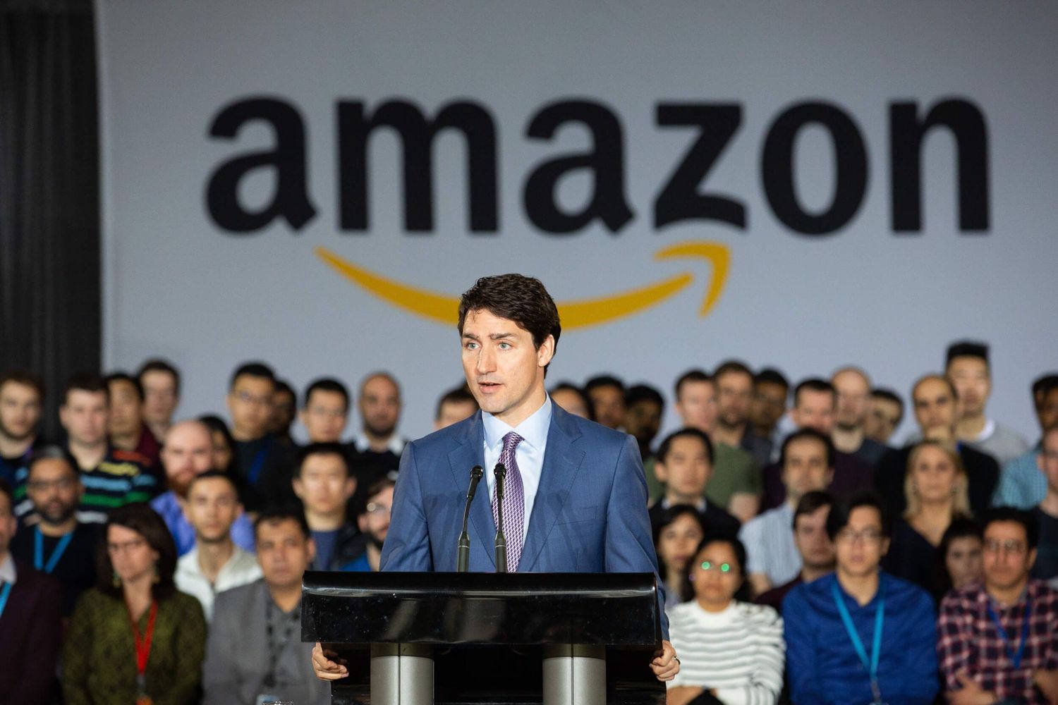 Man speaking at podium in front of Amazon logo with crowd of employees gathered behind him.