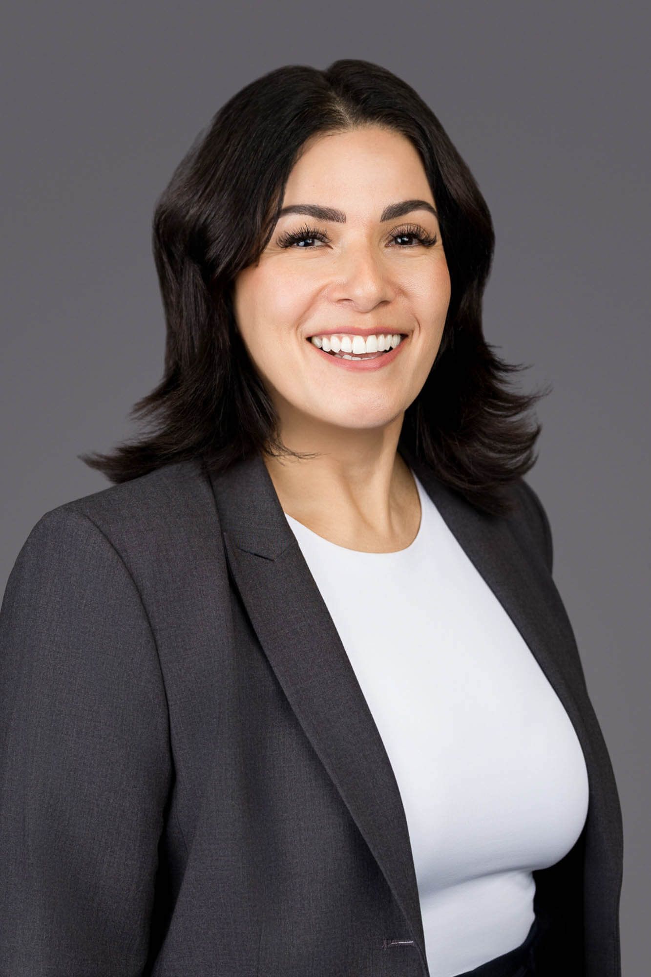 Smiling professional woman with dark hair wearing a white top and gray blazer against a gray background.