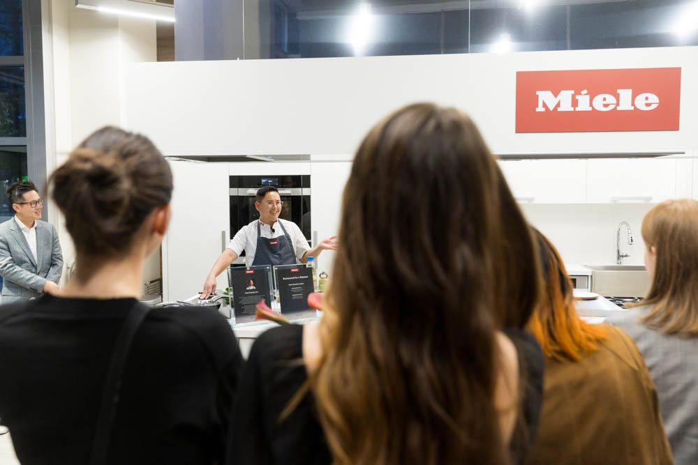Chef demonstrates cooking techniques to an audience at a Miele kitchen appliance showroom event.