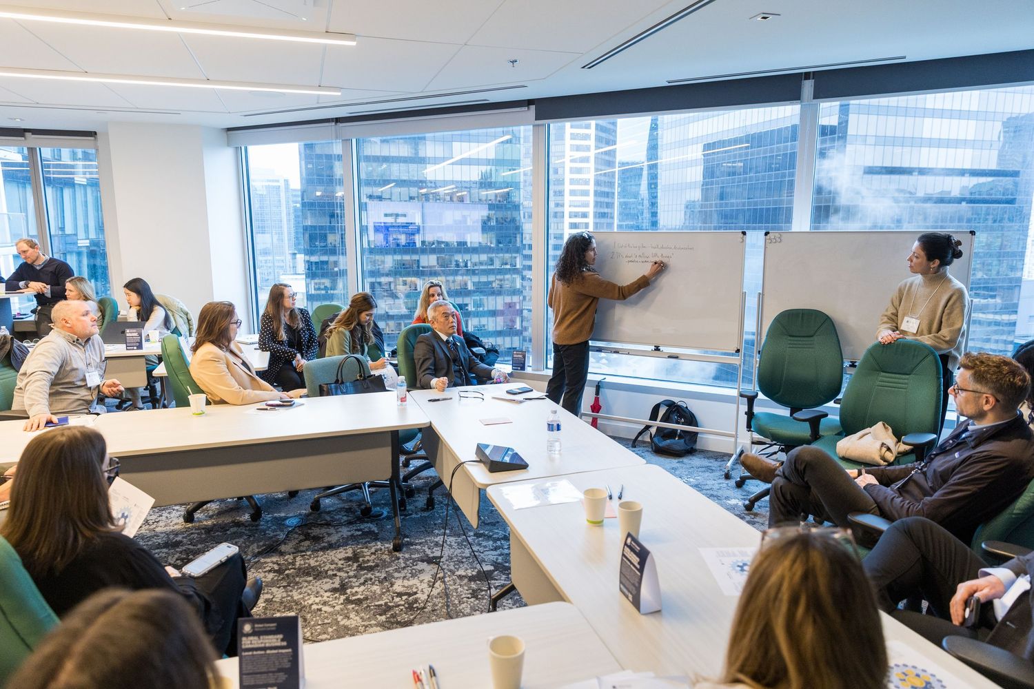 Professionals attend a collaborative workshop in a modern office with floor-to-ceiling windows and a presenter at a whiteboard.
