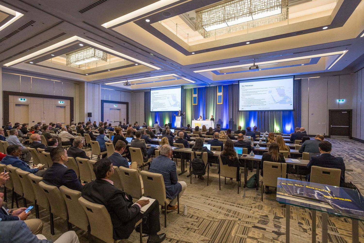 Large conference room filled with attendees watching presentations on dual screens at a professional business summit.