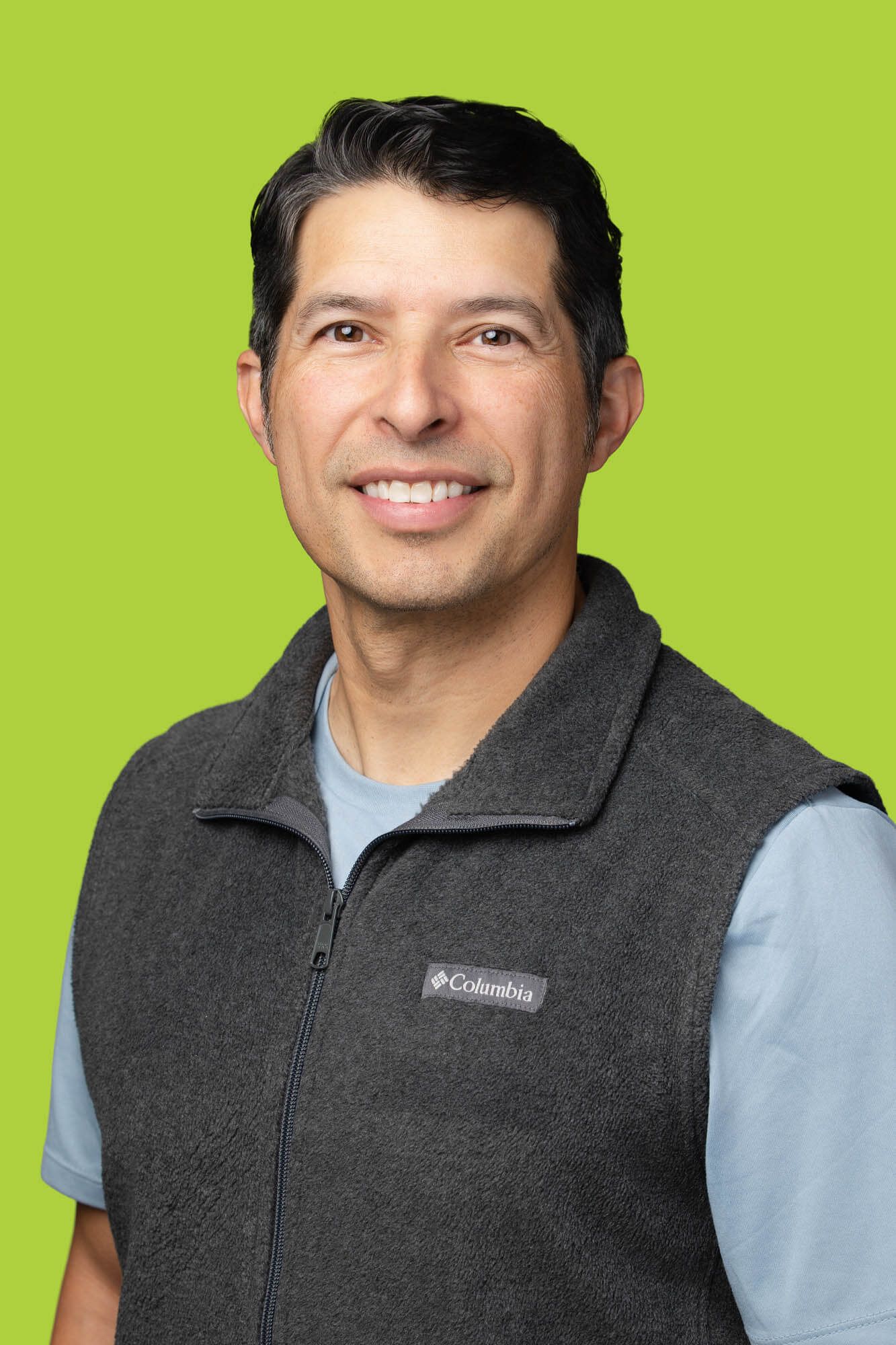 Smiling man in a gray Columbia fleece vest and light blue shirt poses against a bright green background.