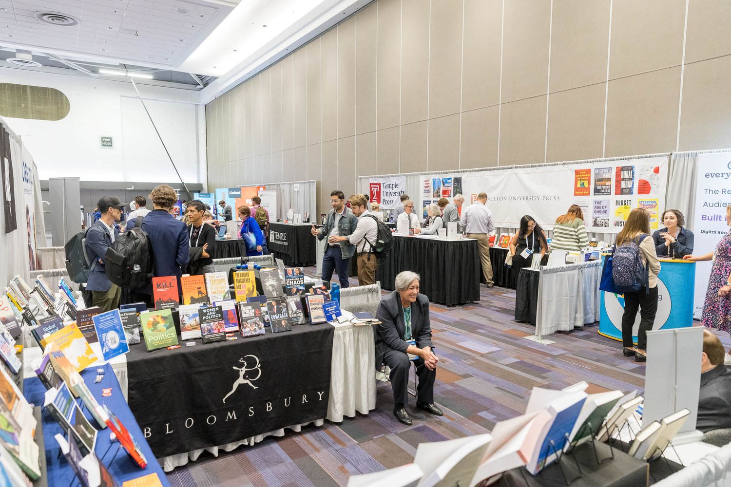 Busy book fair expo hall with Bloomsbury publisher booth displaying colorful books and attendees browsing displays.