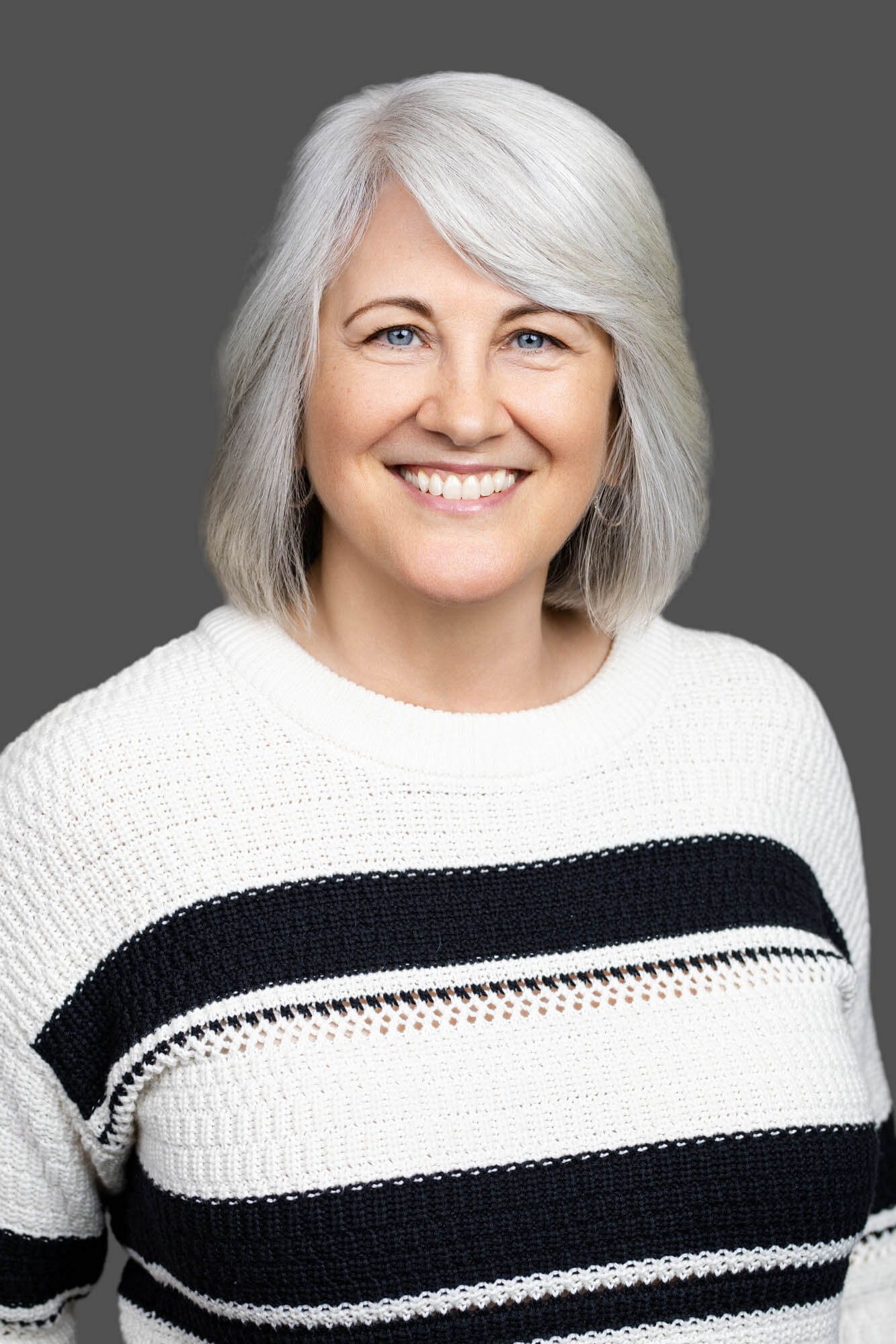 Smiling middle-aged woman with silver bob hair wearing a black and white striped sweater against a gray background.