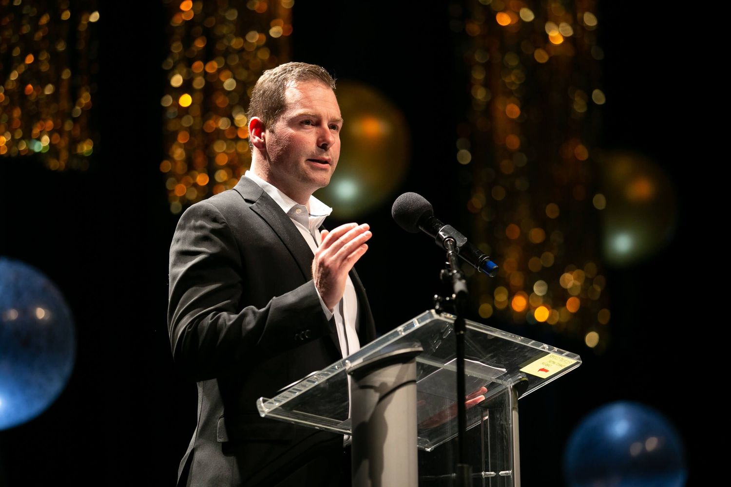 Man in dark suit speaking at podium with microphone, bokeh lights in background at formal event at the River Rock Casino.