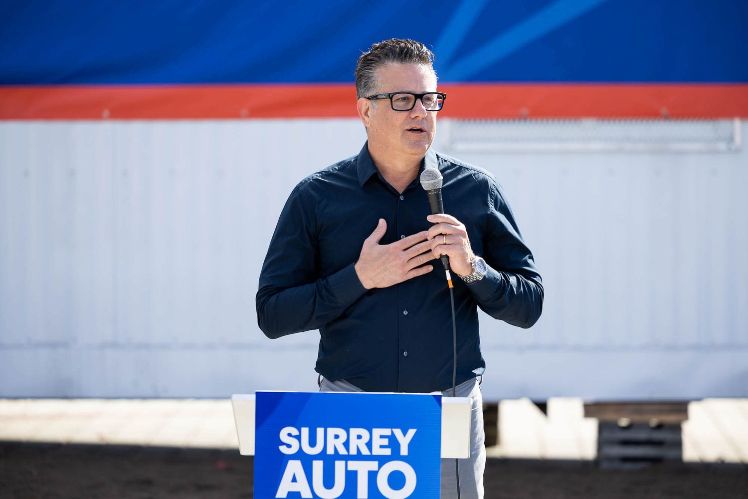 Man speaking at podium with Surrey Auto sign at outdoor event.