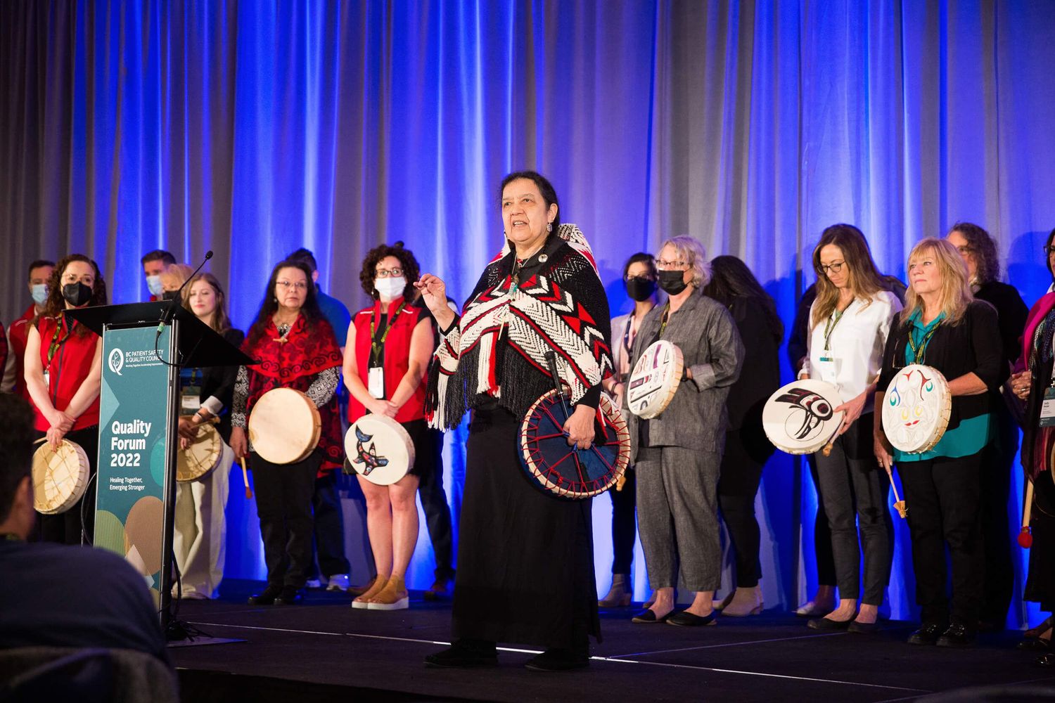 Indigenous woman in traditional regalia leads a drum ceremony at a 2022 Quality Forum conference event at the Hyatt Regency Hotel Vancouver.