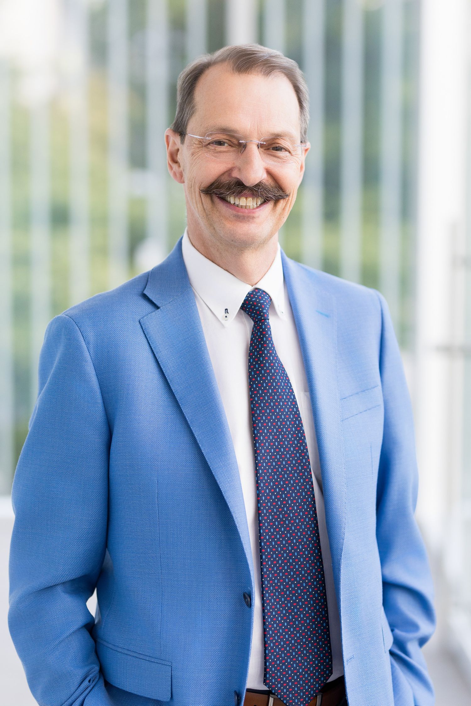 Professional man in blue blazer and tie smiling confidently, wearing glasses and mustache, corporate headshot photo.