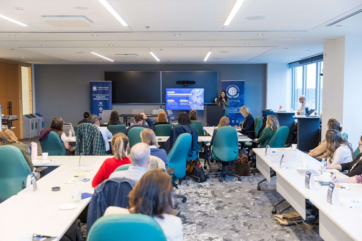Professionals attend a corporate seminar in a modern conference room with teal chairs and a presentation screen.