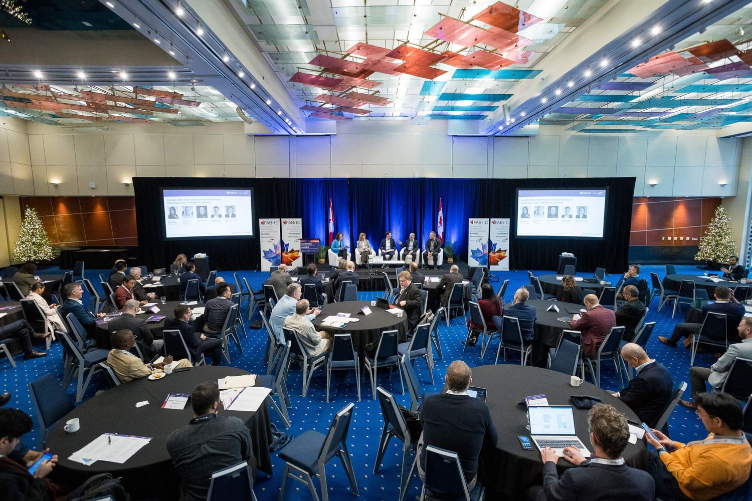 Conference panel discussion in a large ballroom with round tables, attendees, and speakers on a blue-lit stage at the Sheraton Wall Centre.