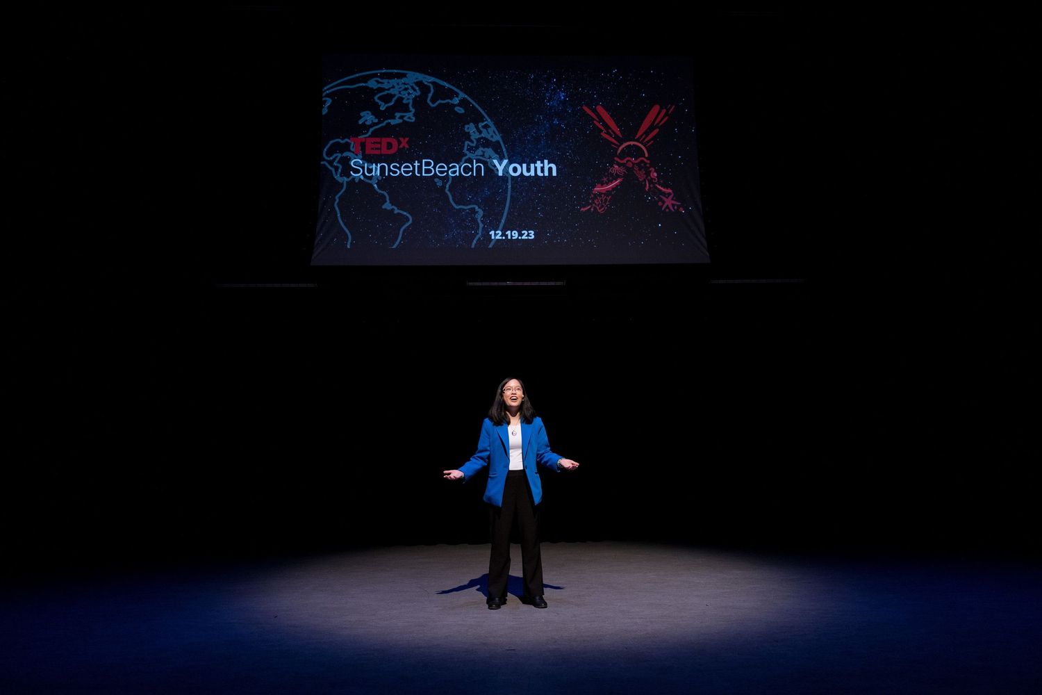 Speaker on stage at TEDx SunsetBeach Youth event, illuminated by spotlight against dark background.