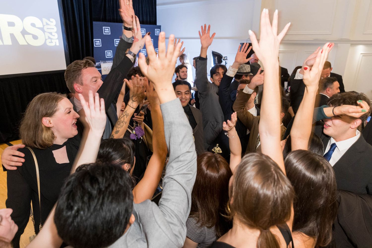 Excited crowd of young professionals raising hands at a corporate celebration event with banner backdrop.