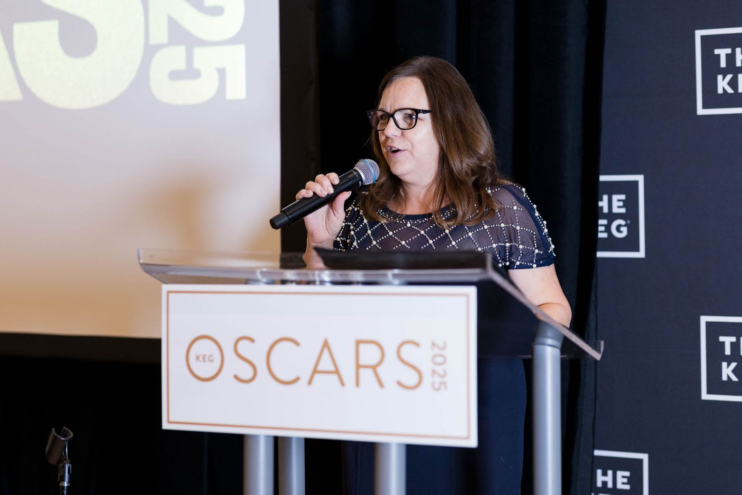 Woman speaking at a podium during the Oscars 2025 awards event at The Keg venue.