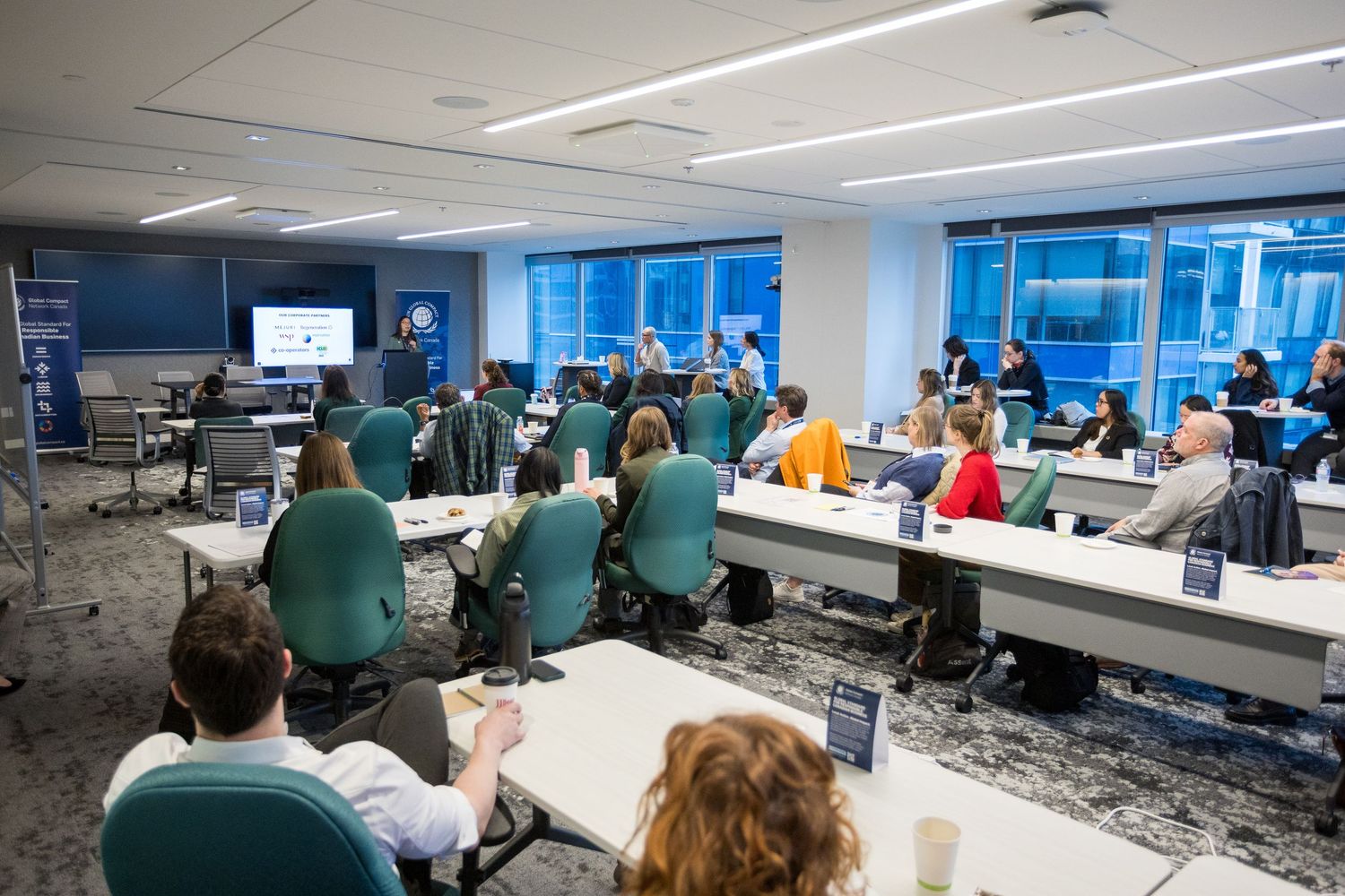 Professionals attend a corporate training seminar in a modern conference room with large windows and rows of tables.
