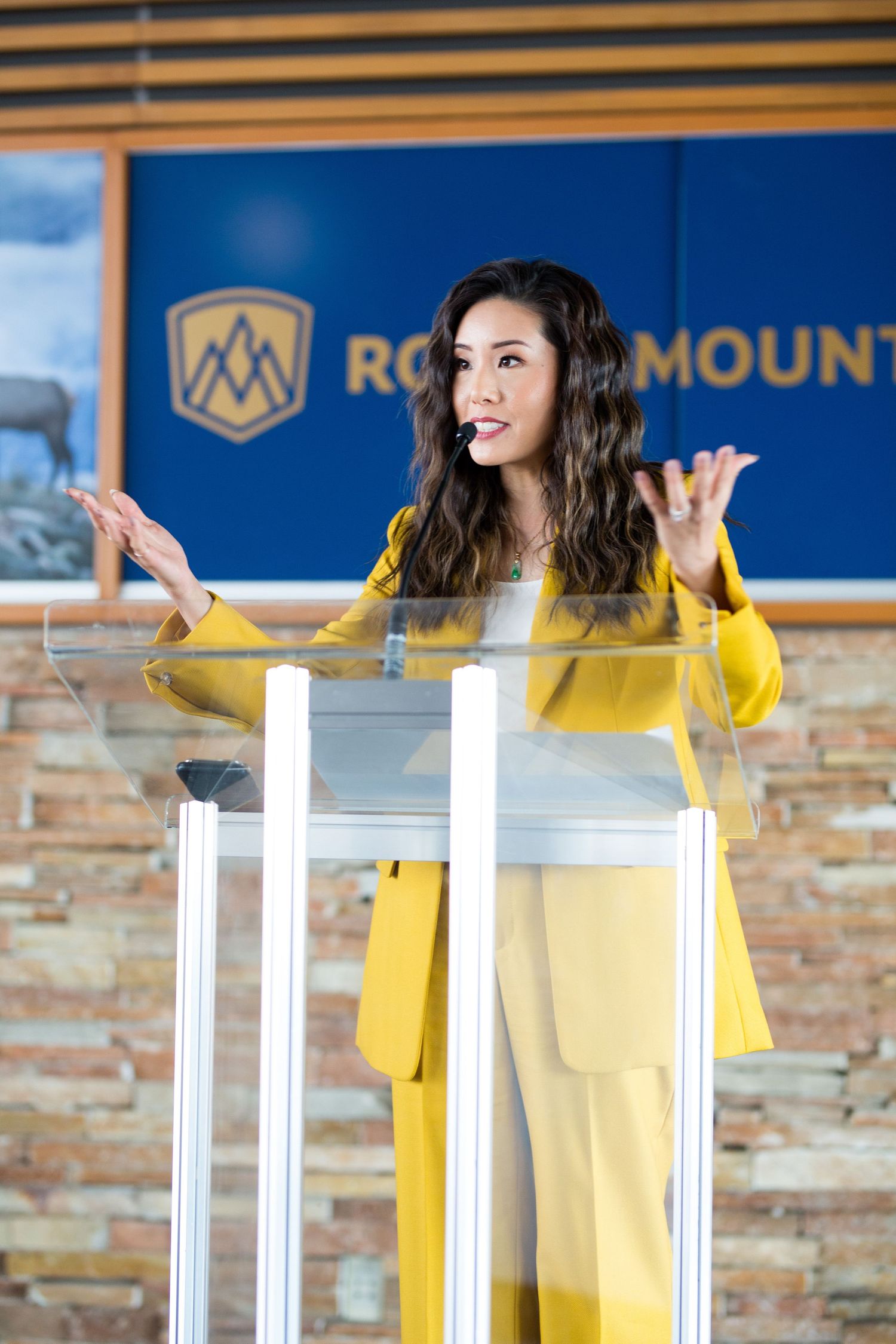 Lien Yeung in yellow suit speaking at a glass podium in front of a Rocky Mountain branded backdrop.