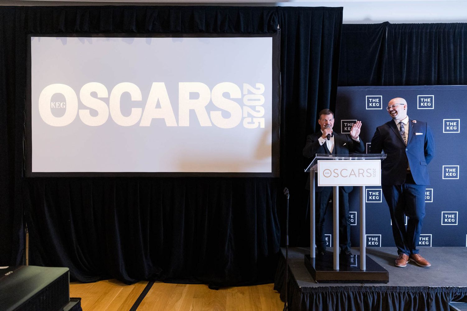 Two men present at the Oscars 2025 event on stage with a large projection screen displaying the Oscars logo.