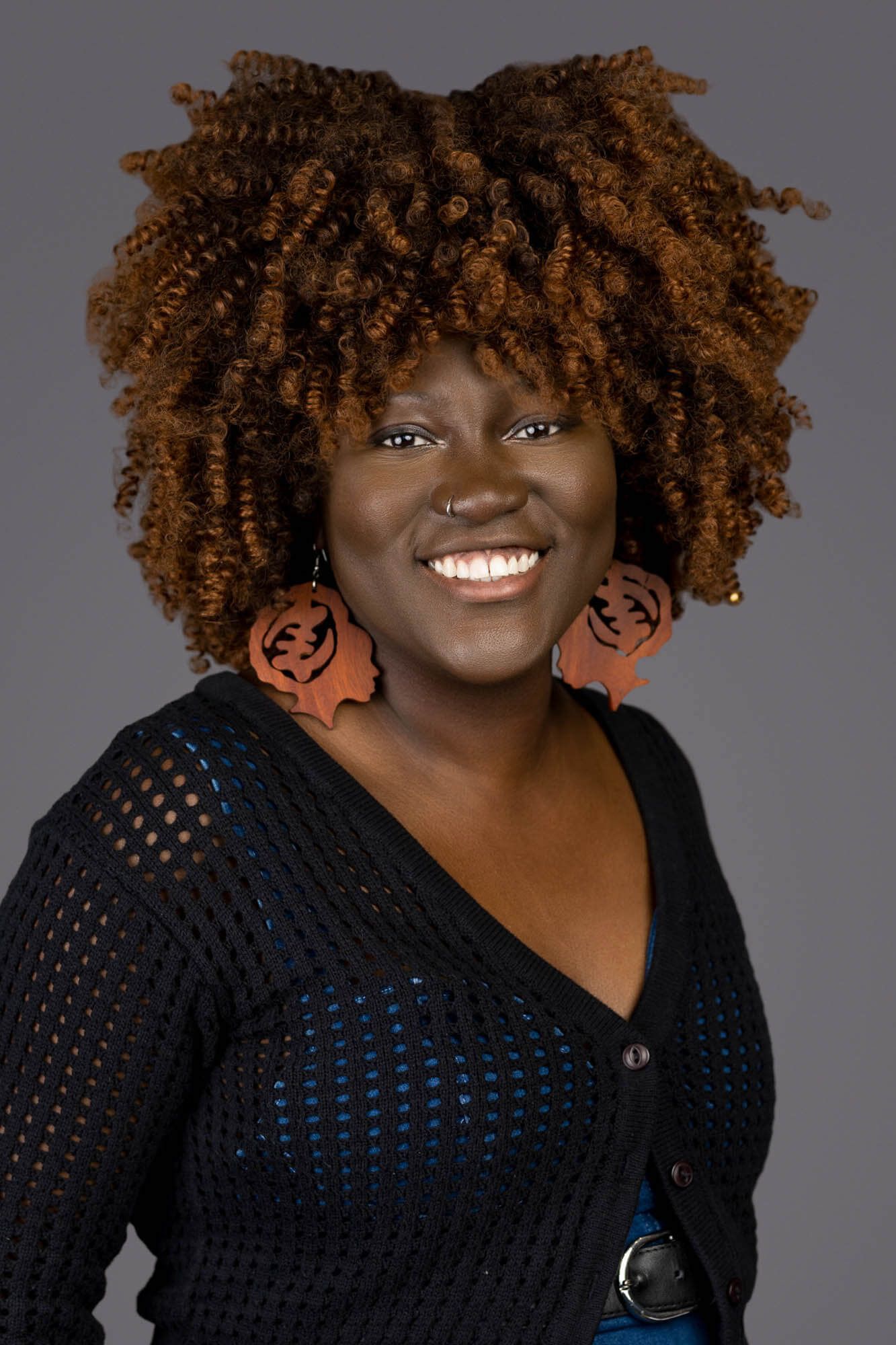 Smiling Black woman with auburn curly hair wearing jack-o-lantern earrings and black knit cardigan against gray background.