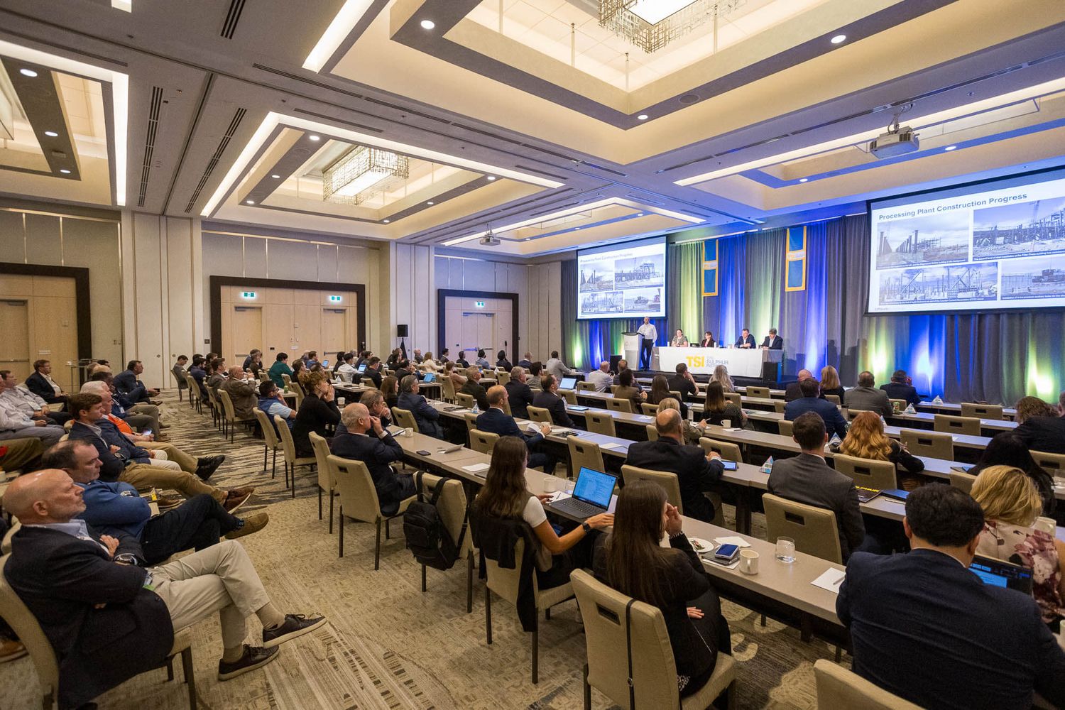 Large conference room filled with attendees seated at rows of tables facing a panel of speakers and large presentation screens.