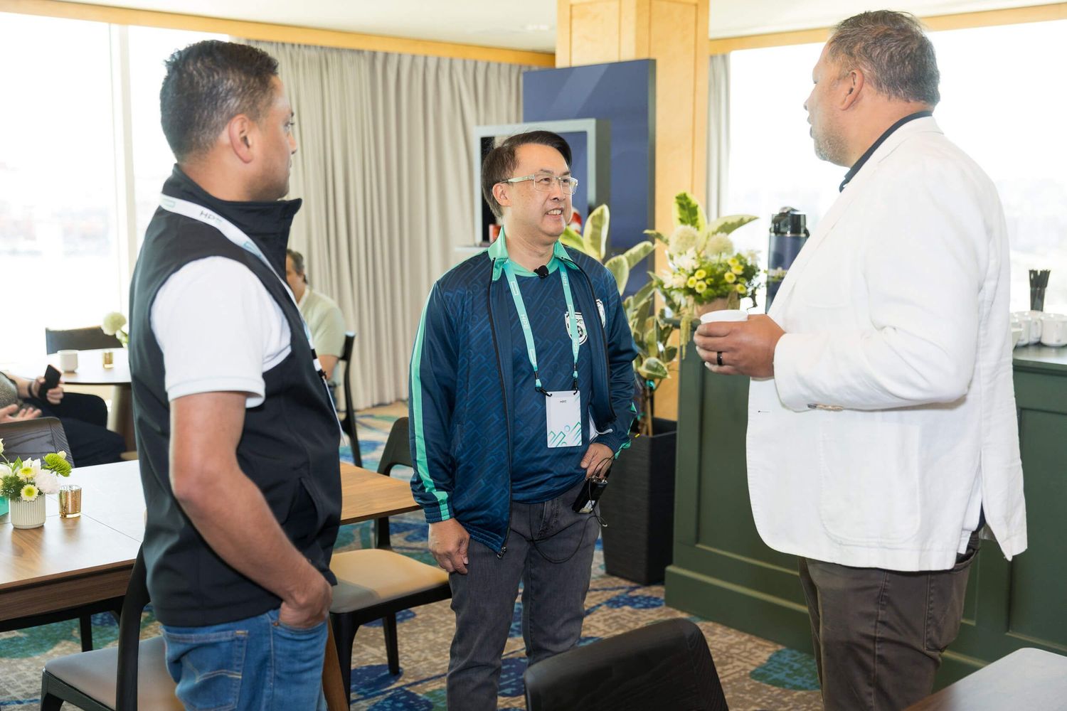 Three men engage in conversation at an indoor networking event with green plants and blue decor.