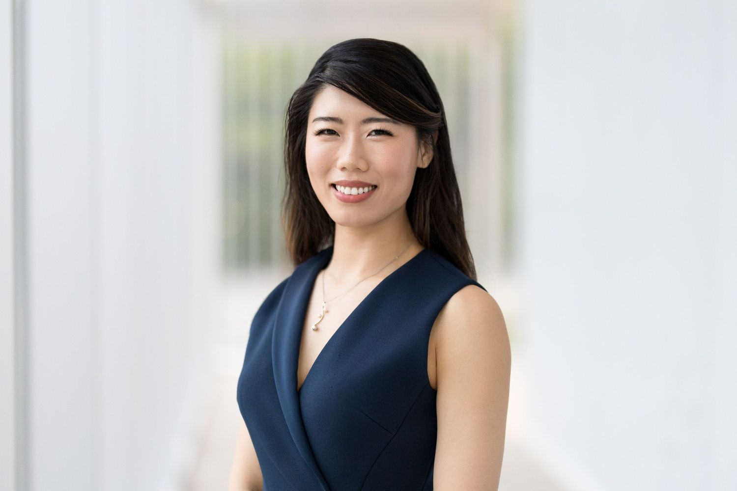 Professional headshot of a smiling young Asian woman in a navy sleeveless dress against a bright white background.