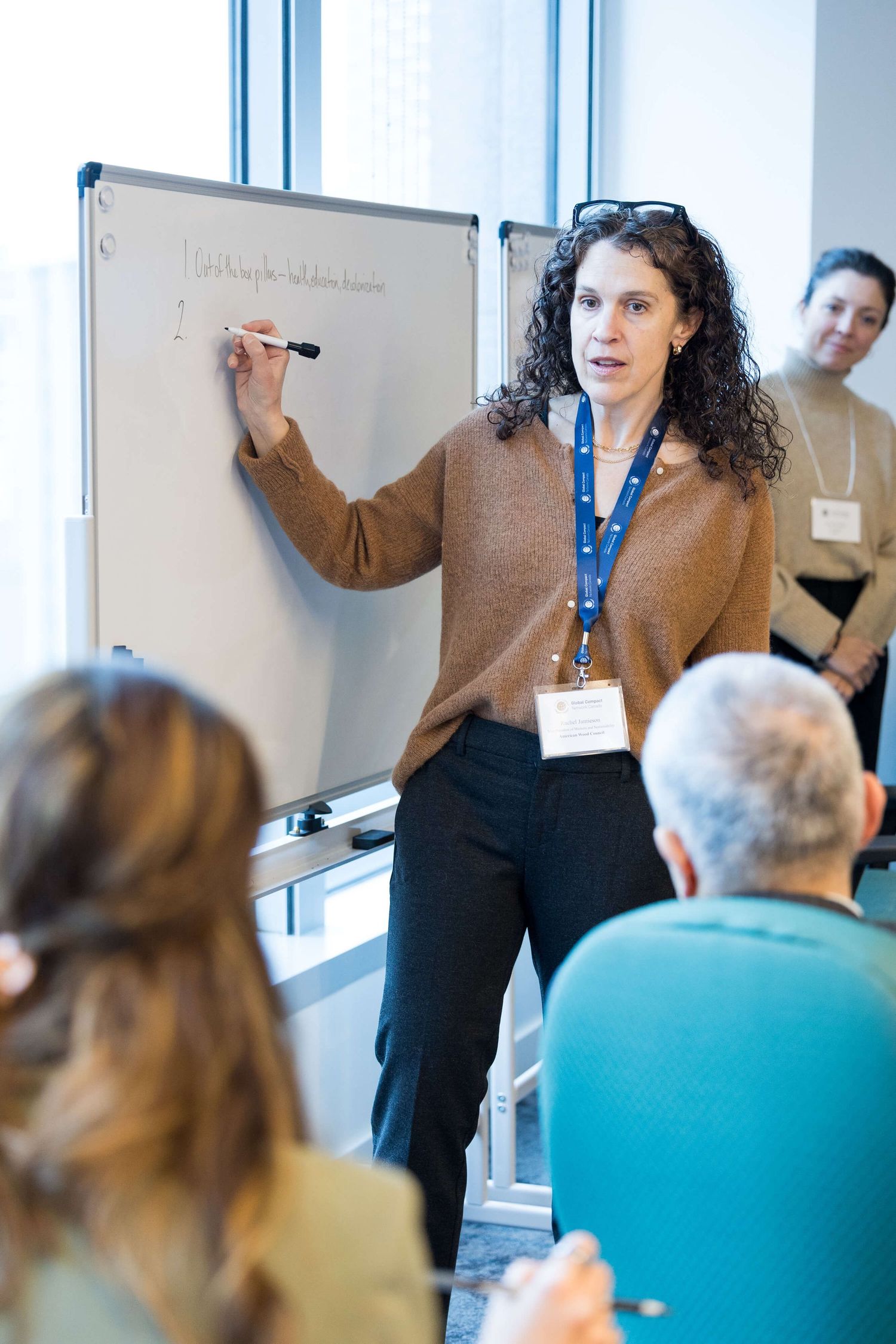 Woman with curly hair presents at whiteboard during professional workshop, wearing lanyard badge and brown sweater.